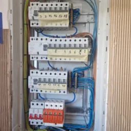 Otwarta skrzynka bezpiecznikowa z zainstalowanymi bezpiecznikami, przewodami w r&oacute;żnych kolorach i oznaczeniami na taśmie maskującej, widoczne elementy instalacji elektrycznej wewnątrz obudowy.