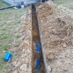 Wiktor Furmańczyk - Wykop w ziemi z niebieską rurą karbowaną na dnie, zabezpieczoną folią, widok z góry, obok domu w budowie.
