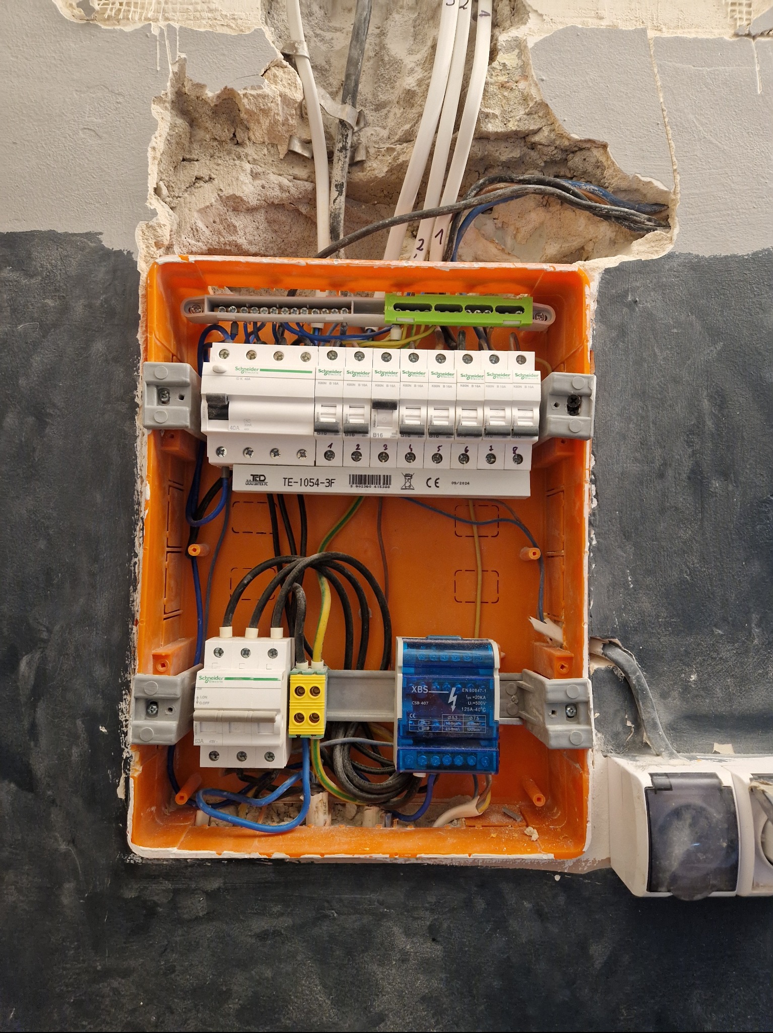 Pomarańczowa skrzynka elektryczna z zamontowanymi bezpiecznikami, widoczne kable i przewody, ściana z widocznymi śladami prac remontowych, częściowo pomalowana na szaro.