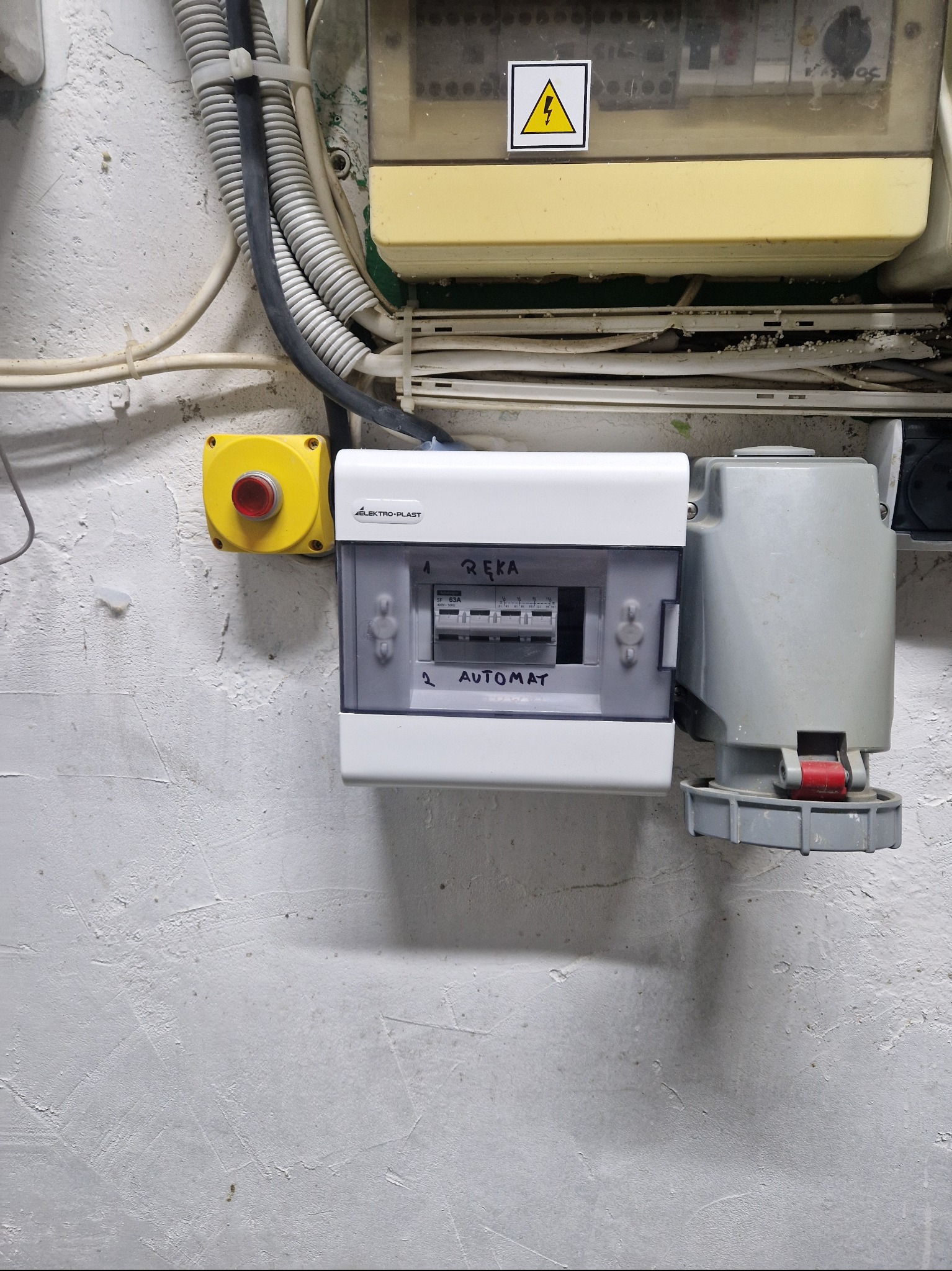 Biała skrzynka bezpiecznikowa marki Elektro-Plast z napisem 'Automat', zamontowana na szarej, chropowatej ścianie obok żółtego przycisku awaryjnego oraz szarego gniazda przemysłowego z czerwoną...