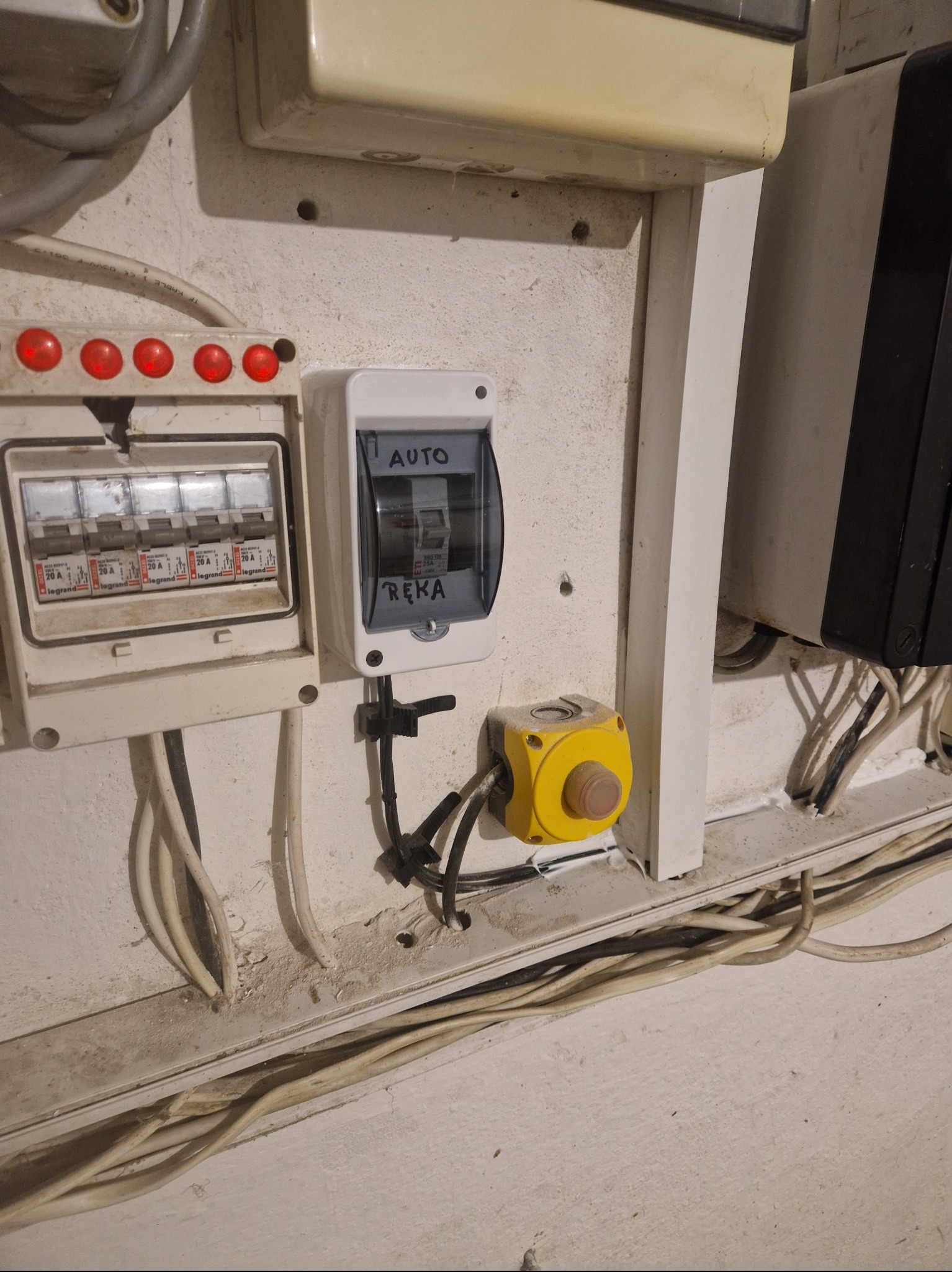 Tablica bezpiecznikowa z wyłącznikami automatycznymi, przycisk awaryjny w żółtej obudowie i widoczne przewody elektryczne na ścianie.
