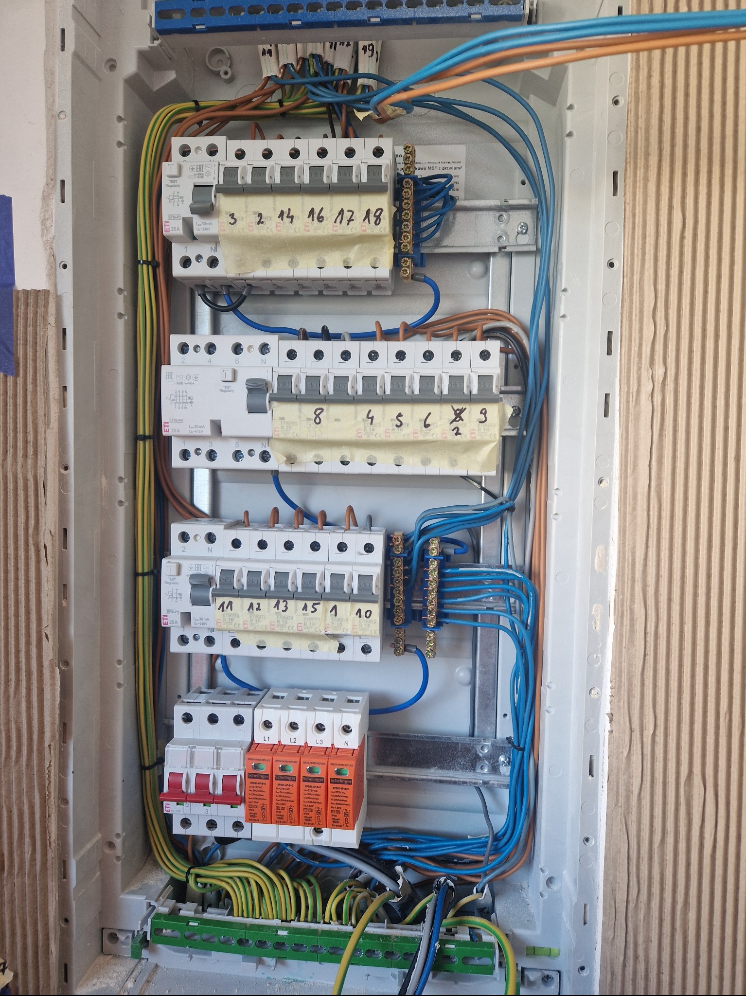 Otwarta skrzynka bezpiecznikowa z zainstalowanymi bezpiecznikami, przewodami w różnych kolorach i oznaczeniami na taśmie maskującej, widoczne elementy instalacji elektrycznej wewnątrz obudowy.