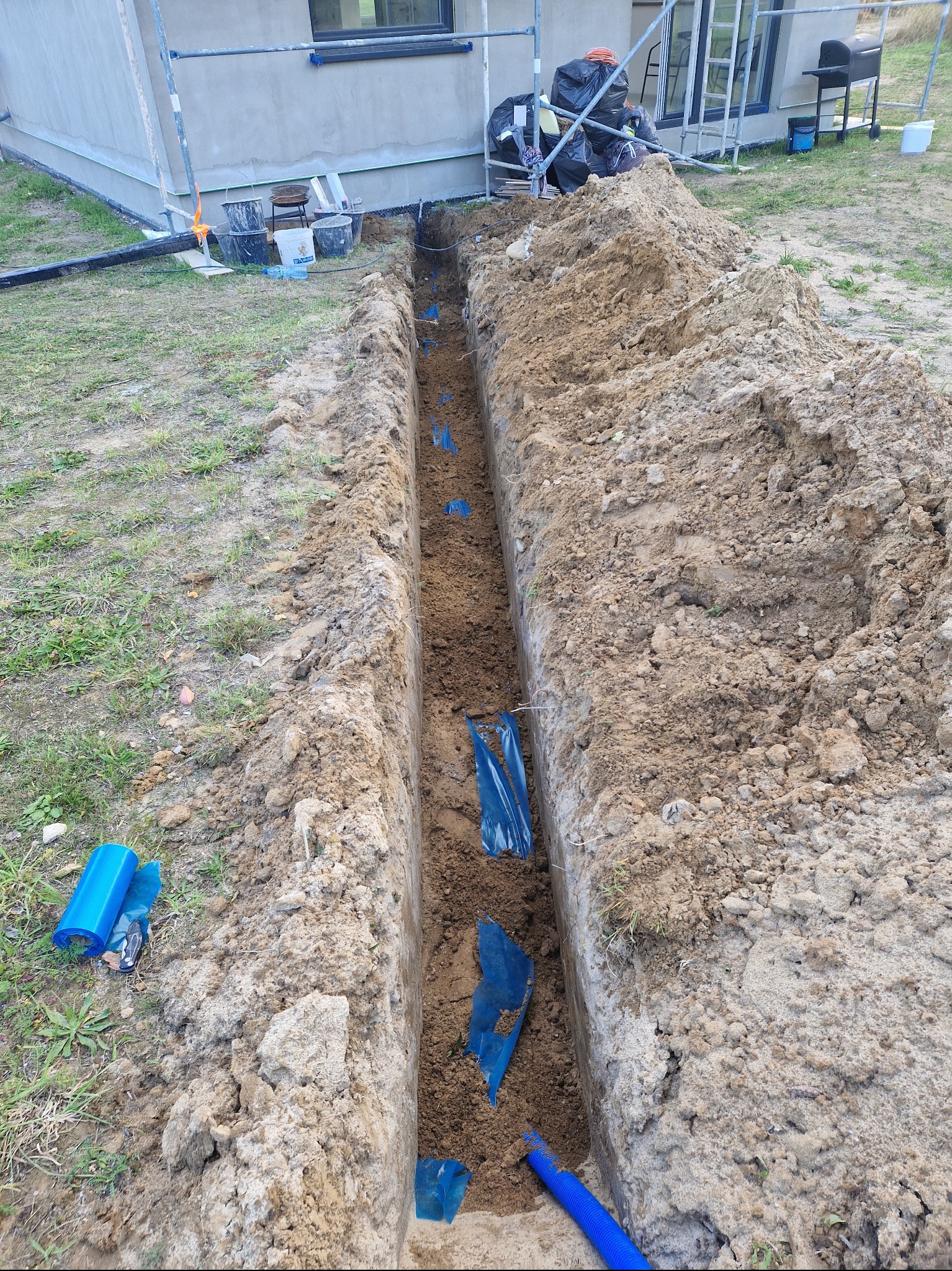 Wykop w ziemi z niebieską rurą karbowaną na dnie, zabezpieczoną folią, widok z góry, obok domu w budowie.