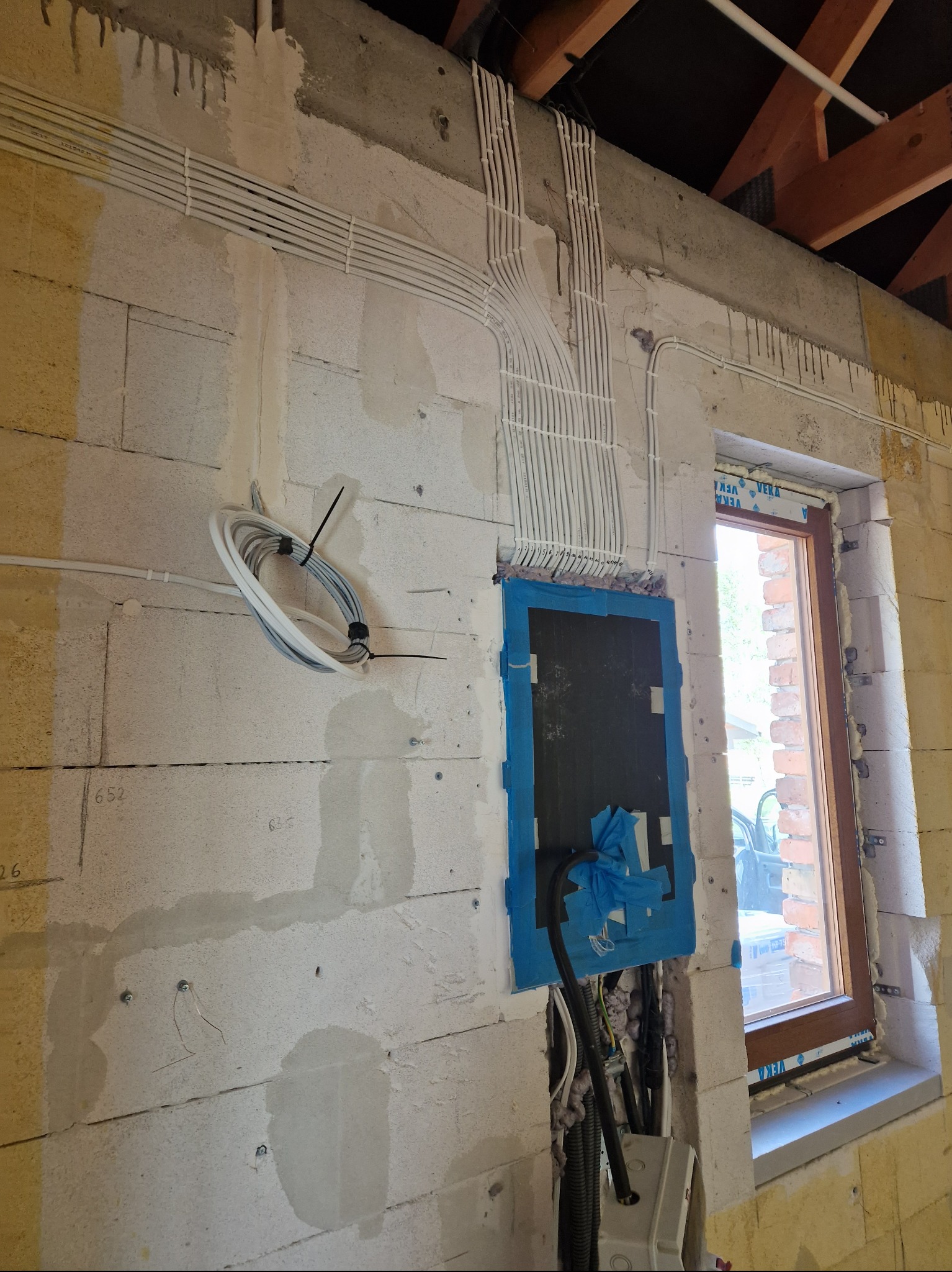 Wewnętrzne okablowanie elektryczne w surowym stanie, białe kable ułożone równolegle na ścianie z betonu komórkowego, zabezpieczona taśmą rozdzielnica, widoczne okno z brązową ramą.