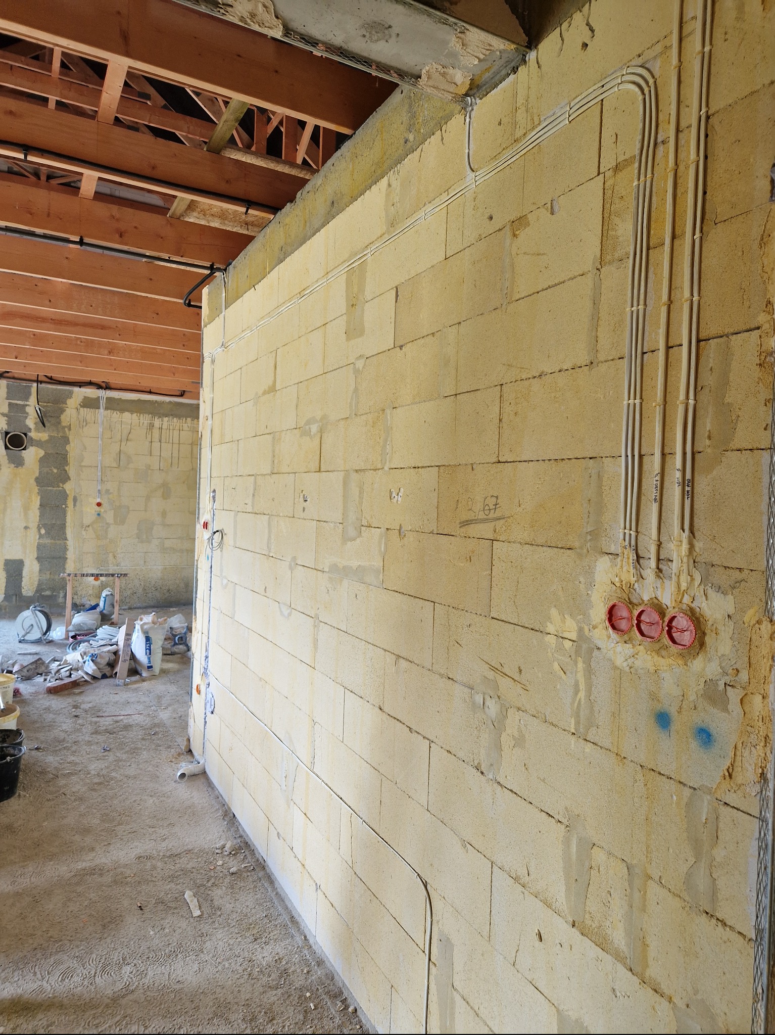 Ściana z betonu komórkowego z poprowadzonymi przewodami elektrycznymi w rurkach oraz puszkami pod gniazdka, widoczne także elementy konstrukcji stropu i podłoga w stanie surowym.