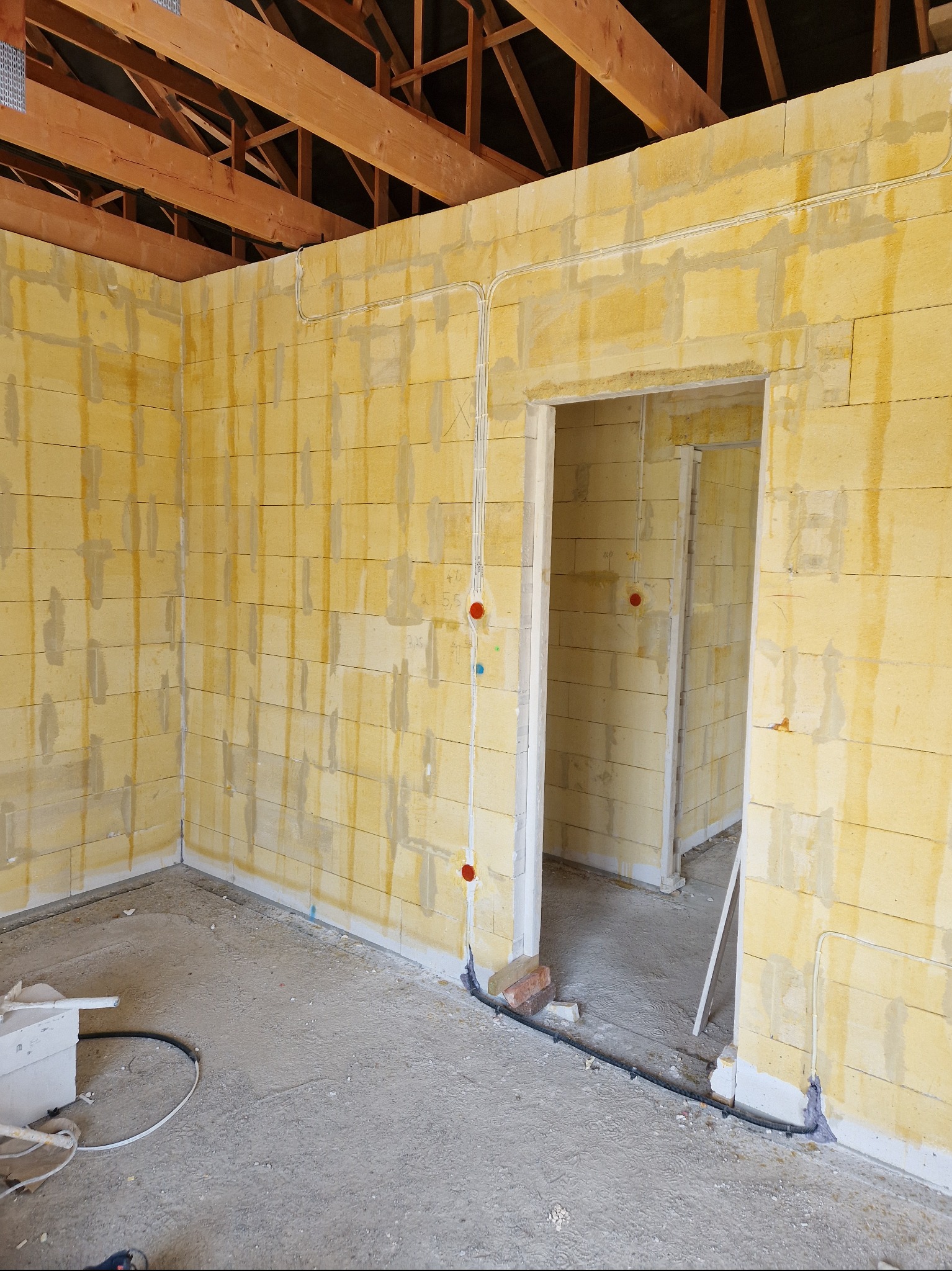 Wnętrze budynku w trakcie prac instalacyjnych; widoczne ściany z bloczków, strop drewniany, przewody elektryczne poprowadzone po ścianach, puszki instalacyjne w kolorze czerwonym i niebieskim,...