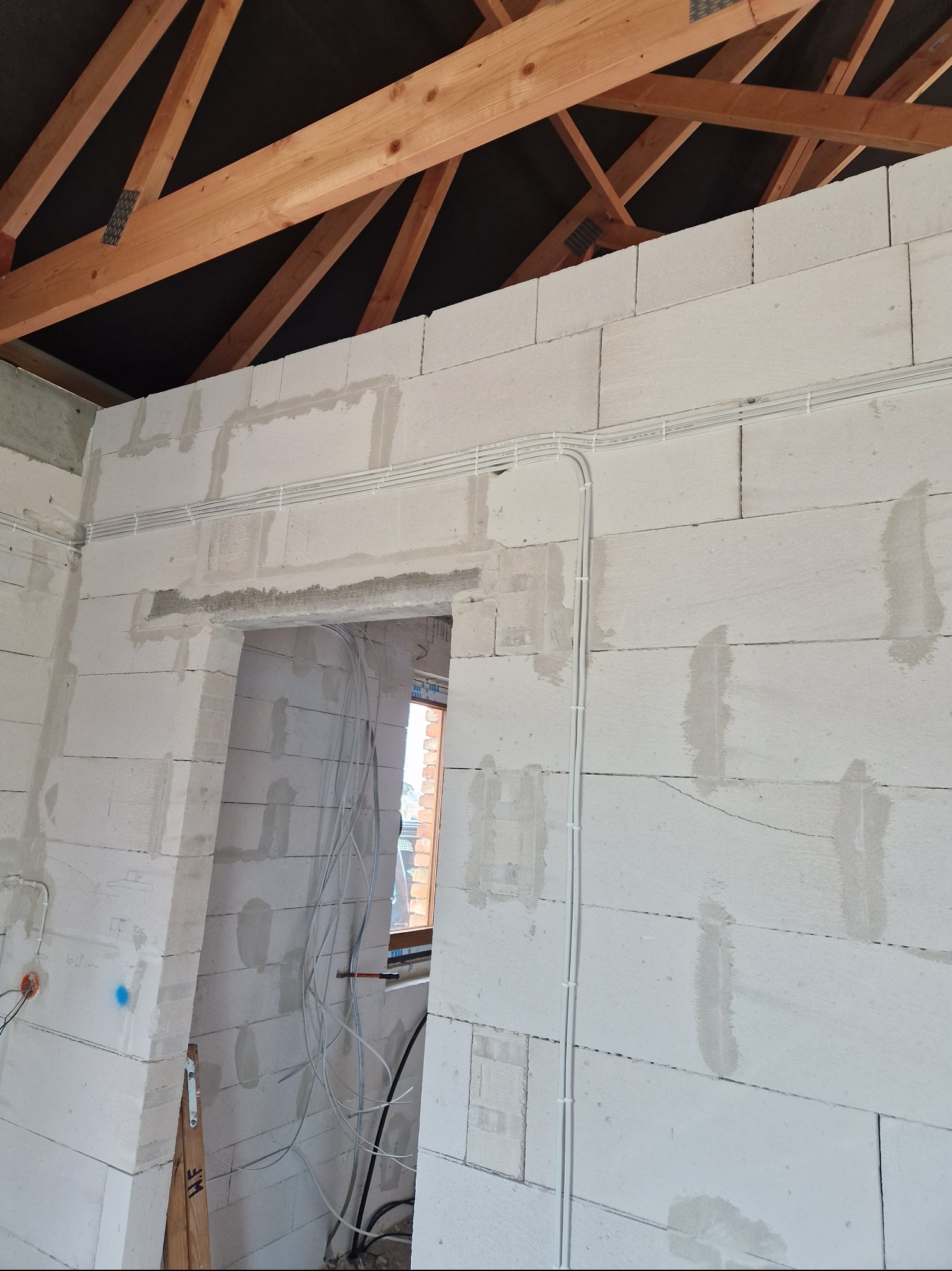 Wnętrze w stanie surowym z widocznymi przewodami elektrycznymi zamocowanymi do ściany z betonu komórkowego, drewnianą konstrukcją dachu i otworami na okna i drzwi.