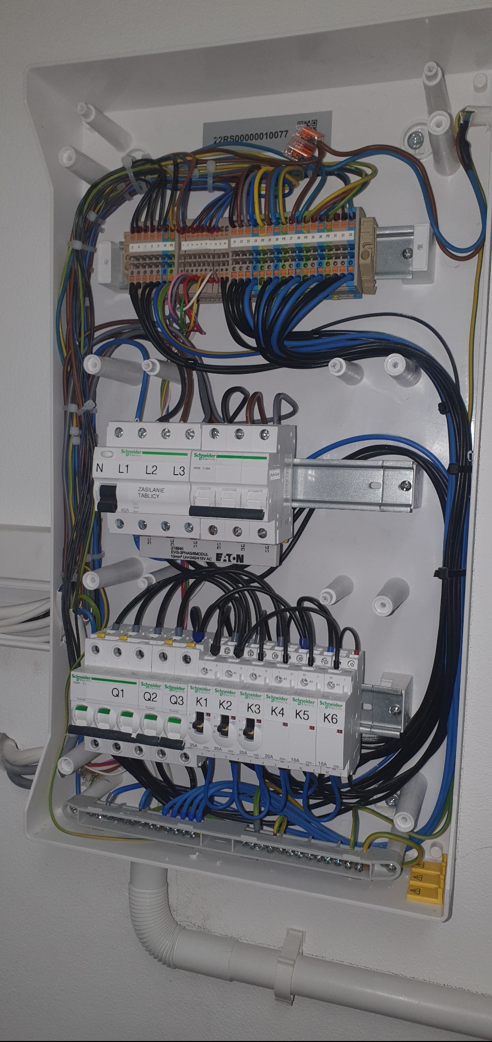 Otwarta skrzynka elektryczna z zainstalowanymi bezpiecznikami, przewodami różnej grubości i koloru oraz złączkami, widoczne elementy firm Schneider i Eaton, dolny przewód instalacyjny z białego PCV