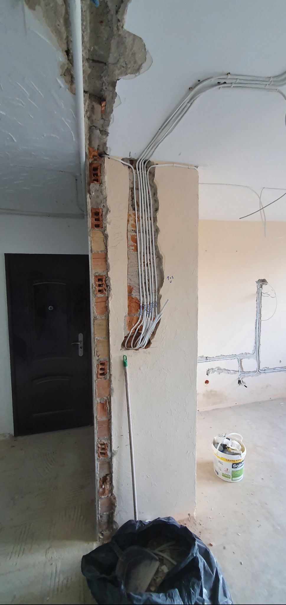 Białe przewody elektryczne ułożone w bruździe w ścianie z cegły, widoczne fragmenty nieotynkowanej ściany, w tle czarne drzwi i wiadro z resztkami farby.