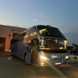 Szary autokar GTV BUS zaparkowany na brukowanym placu, z otwartymi drzwiami i włączonymi światłami, sfotografowany o zmierzchu na tle budynku i zachmurzonego nieba.