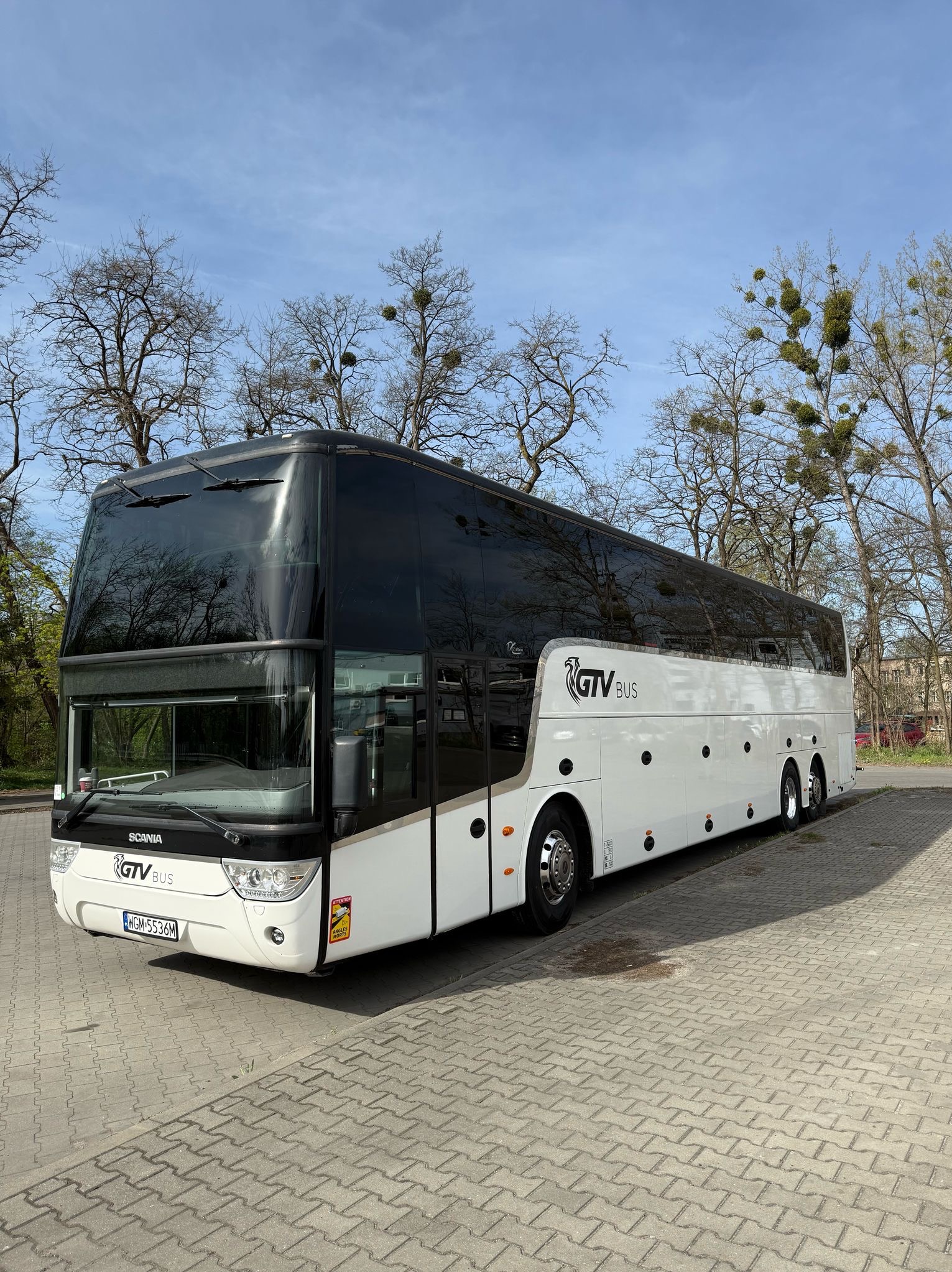 Nowoczesny, biały autokar turystyczny Scania z czarnymi szybami, zaparkowany na brukowanym placu, z logo 'GTV BUS' na karoserii i tablicą rejestracyjną WGM 5536M.