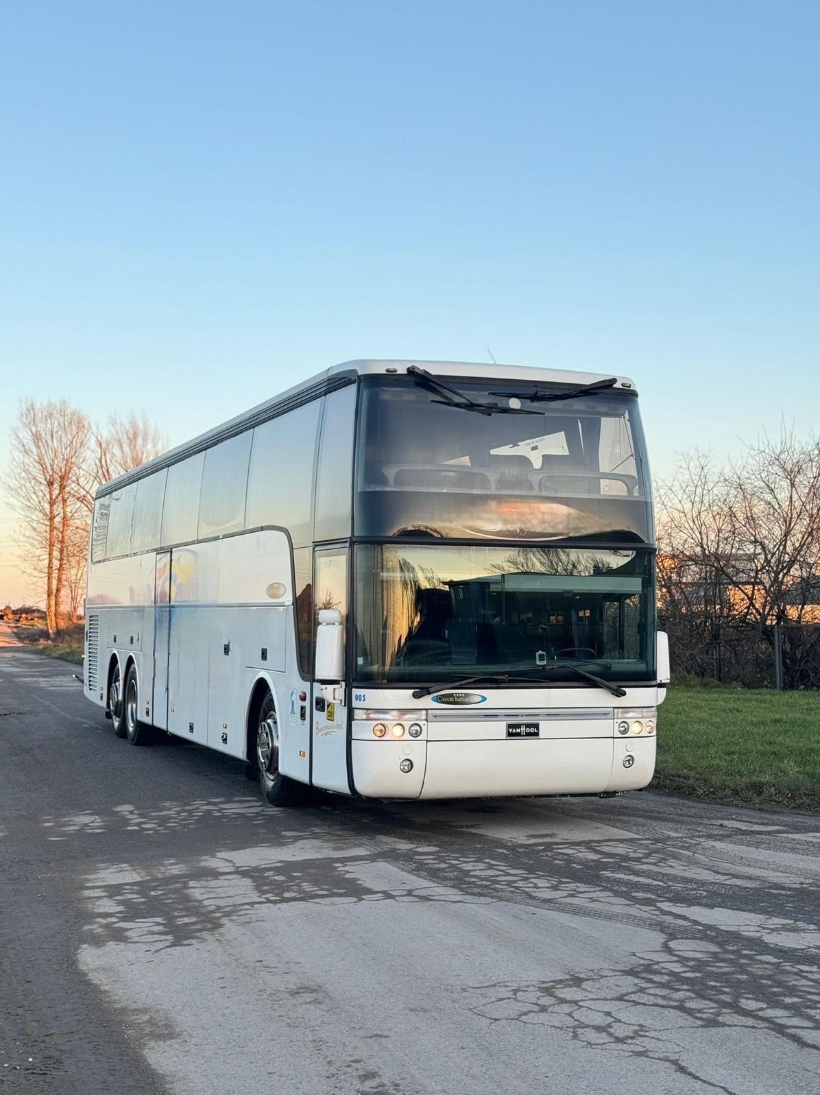 Dwupokładowy, biały autokar turystyczny Van Hool na szosie, widok z przodu, w tle drzewa i błękitne niebo.