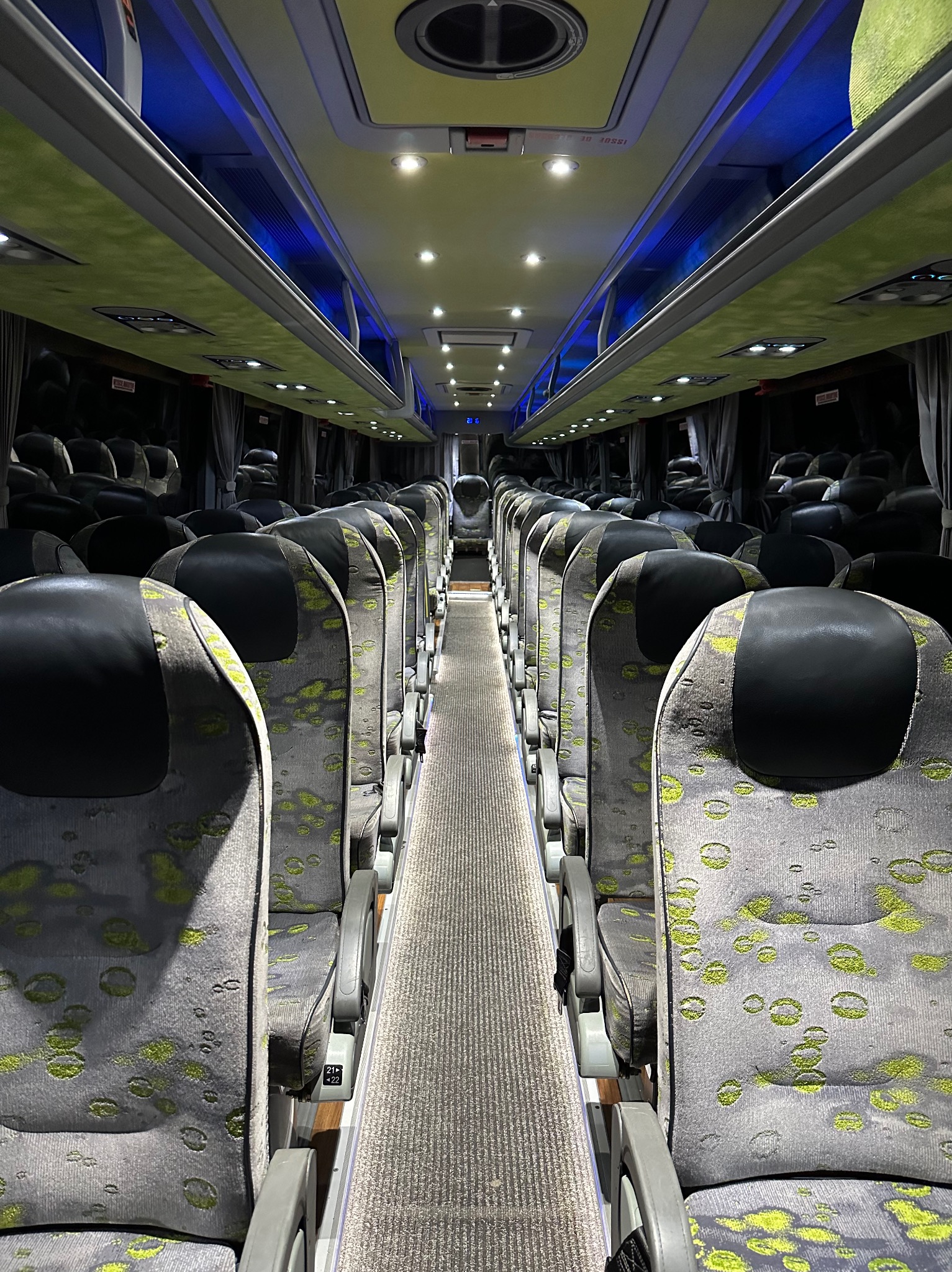 Wnętrze autobusu z rzędami szarych foteli z zielonymi wzorami, czarnymi zagłówkami i centralnym przejściem z szarą wykładziną, oświetlone punktowymi lampami i niebieskim światłem w górnej części.
