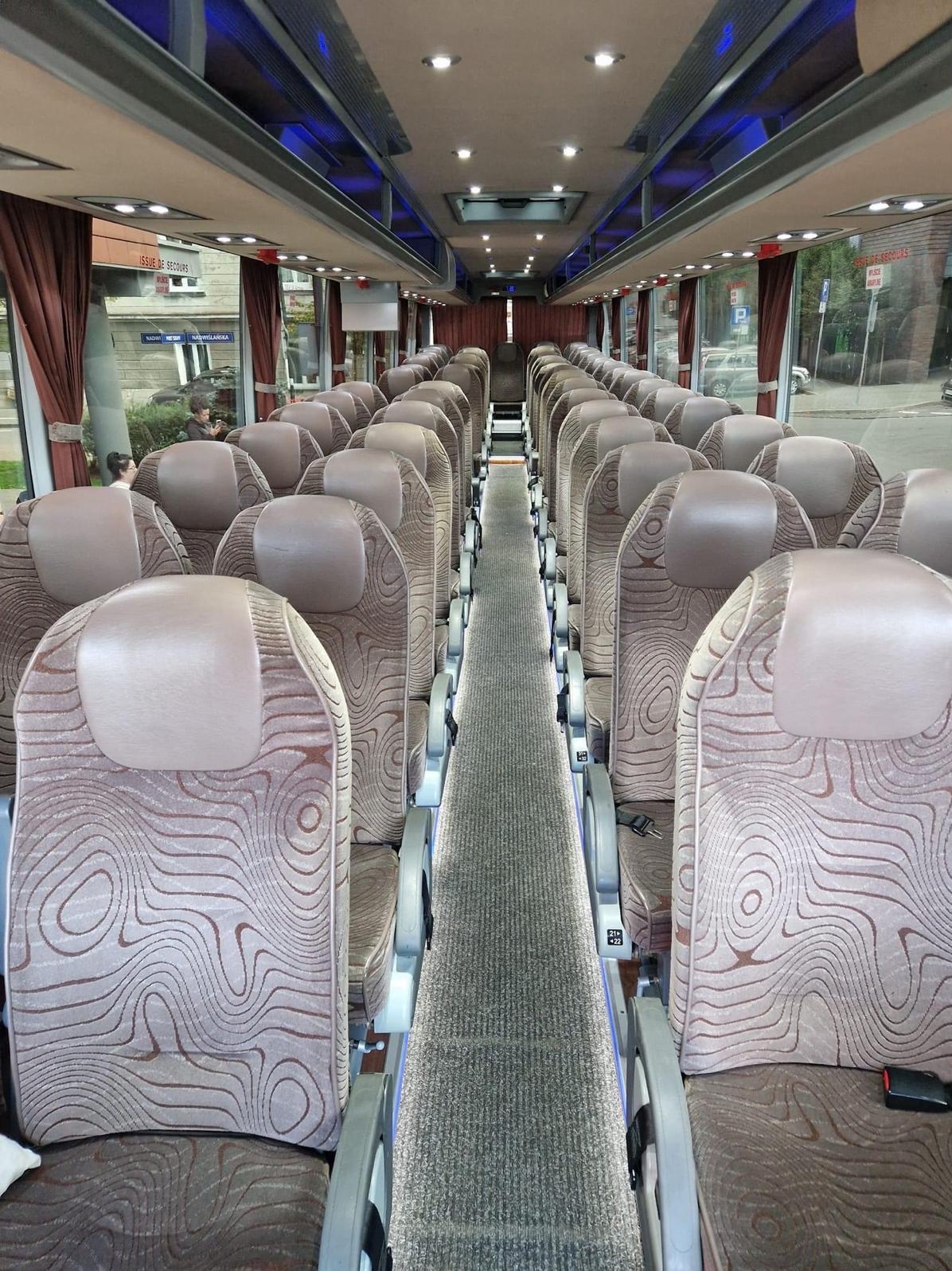 Wnętrze autobusu turystycznego z rzędami brązowo-beżowych foteli z abstrakcyjnym wzorem, wąski dywanik na środku, widok z przodu pojazdu.