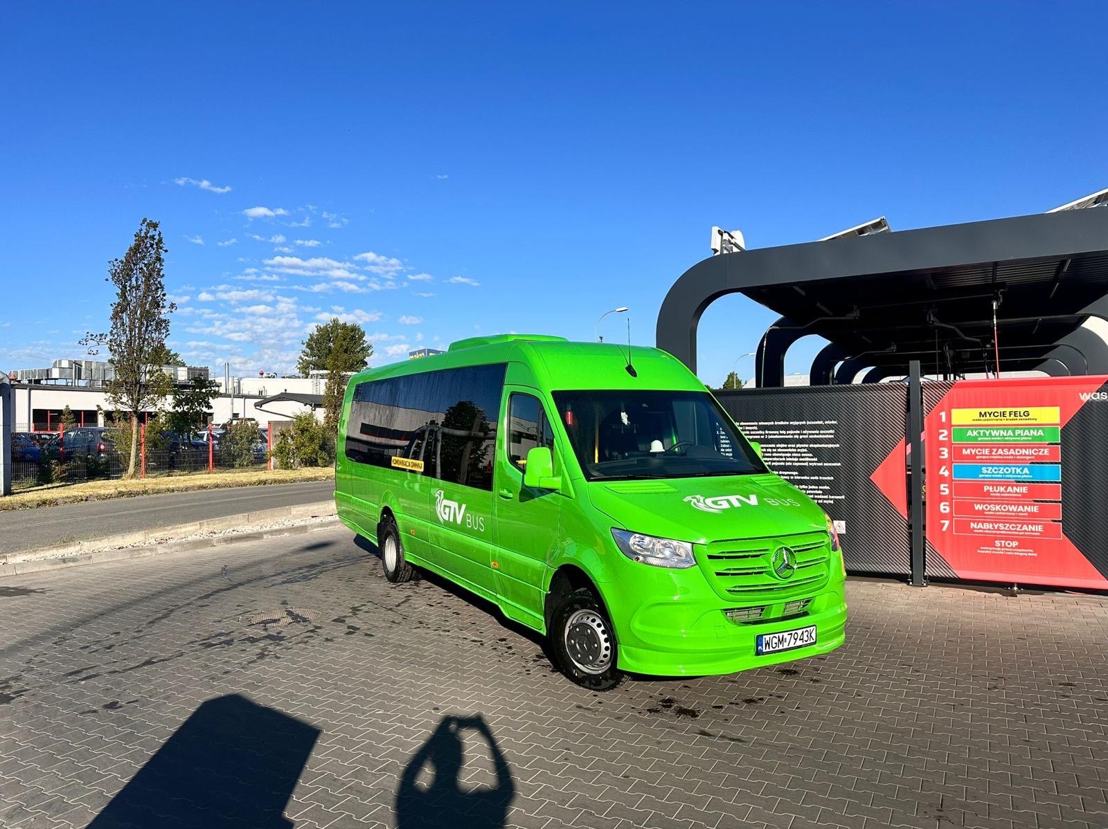 Jasnozielony bus marki Mercedes z logo 'GTV BUS' zaparkowany na kostce brukowej przed automatyczną myjnią samochodową w słoneczny dzień.