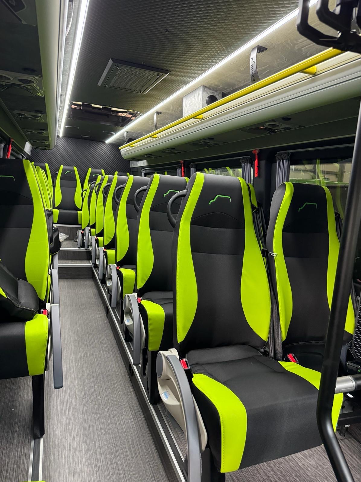Wnętrze autobusu z rzędami czarno-zielonych foteli, szarą podłogą i oświetleniem LED, widok z perspektywy pasażera.