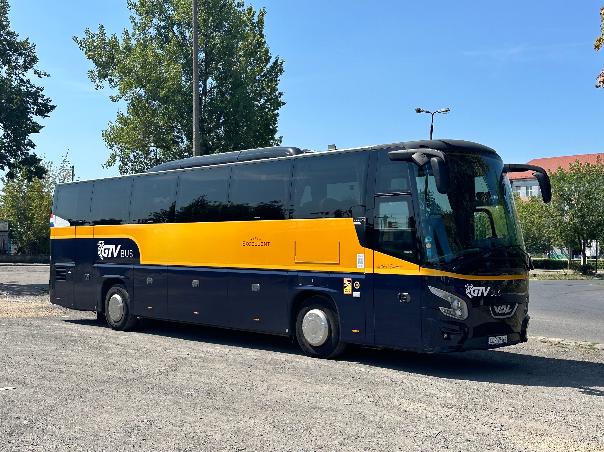 Autokar turystyczny GTV Bus z żółtym pasem na ciemnym tle, zaparkowany na żwirowym parkingu w słoneczny dzień.