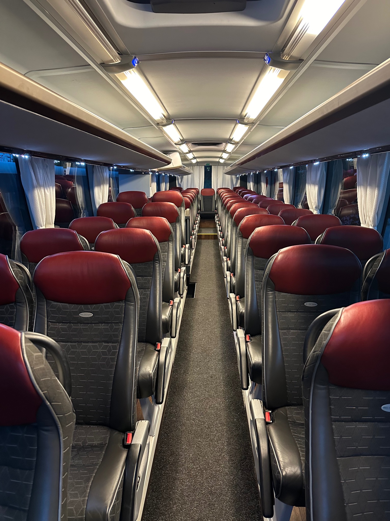 Wnętrze autobusu z rzędami foteli z czerwonymi zagłówkami, oświetlonych podłużnymi lampami, z widocznymi zasłonkami w oknach.