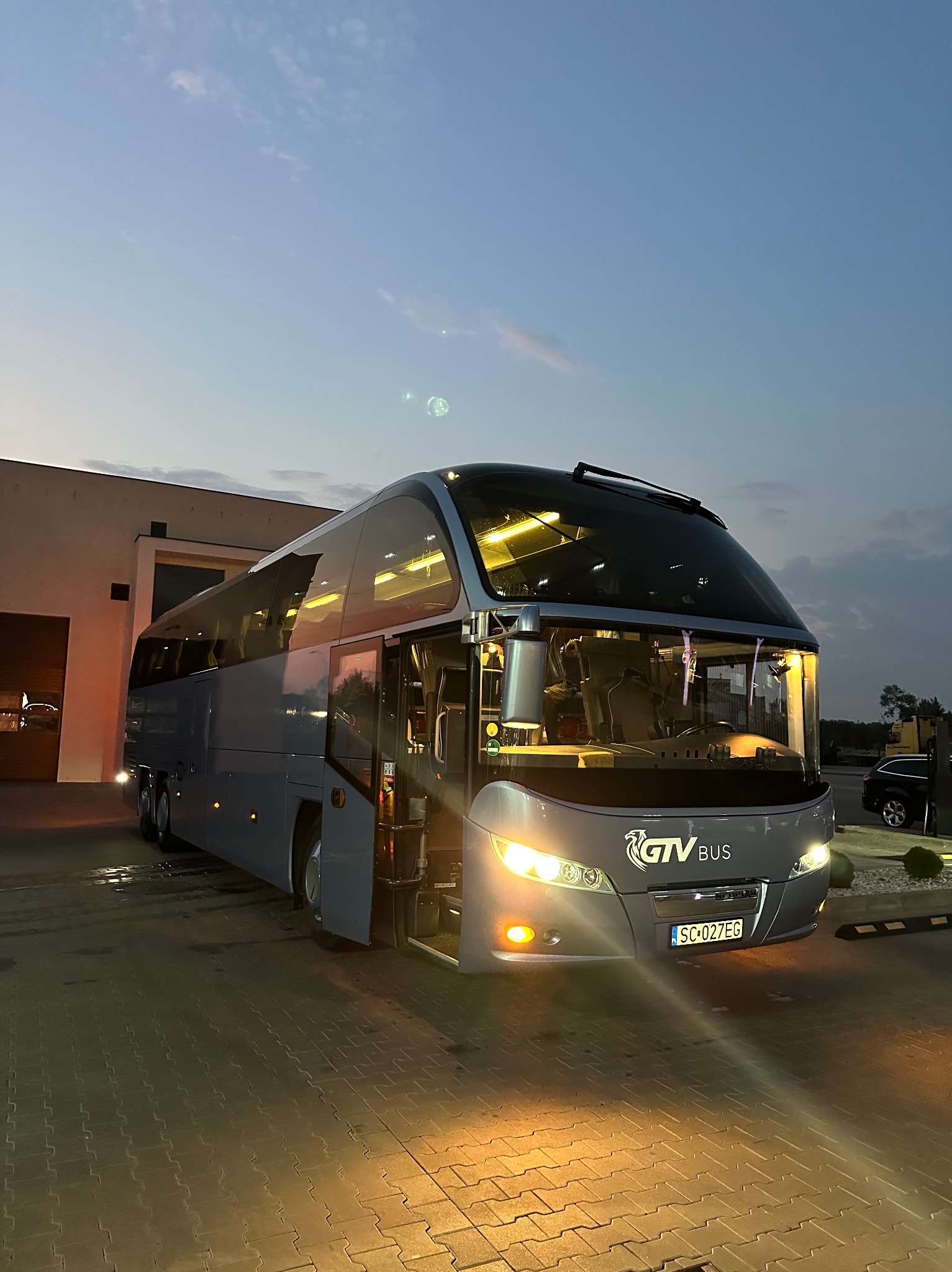 Szary autokar GTV BUS zaparkowany na brukowanym placu, z otwartymi drzwiami i włączonymi światłami, sfotografowany o zmierzchu na tle budynku i zachmurzonego nieba.