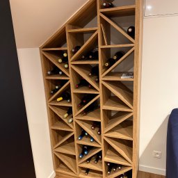 AM Deko kuchnie - Drewniany regał na wino, wkomponowany w skos dachu, z butelkami wina w trójkątnych przegródkach. Jasne drewno, nowoczesny design, oświetlenie punktowe.