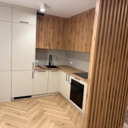 Nowoczesna kuchnia z białymi szafkami i drewnianymi akcentami, parkiet w jodełkę, pionowe lamele dekoracyjne i czarna armatura. Minimalistyczny design.