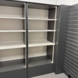 Szare, podświetlane półki z białymi wnękami, część ekspozycji meblowej z panelami ściennymi na metalowe zawieszki, widok z dołu.