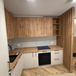 Nowoczesna kuchnia z drewnianymi szafkami i białymi frontami, wykończona drewnianą ścianką działową. Parkiet w jodełkę, czarny zlew i piekarnik.