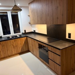 AM Deko - Nowoczesna kuchnia na wymiar z drewnianymi frontami i czarnym blatem w Jarosławiu. Widoczna płyta indukcyjna i piekarnik. Minimalistyczny design, ciepłe oświetlenie.