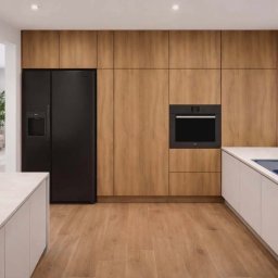 MARIUSZ LEWKIEWICZ - Nowoczesna kuchnia z drewnianymi frontami szafek, czarną lodówką i piekarnikiem. Biały blat i niebieski zlew. Widok na salon z beżową sofą.