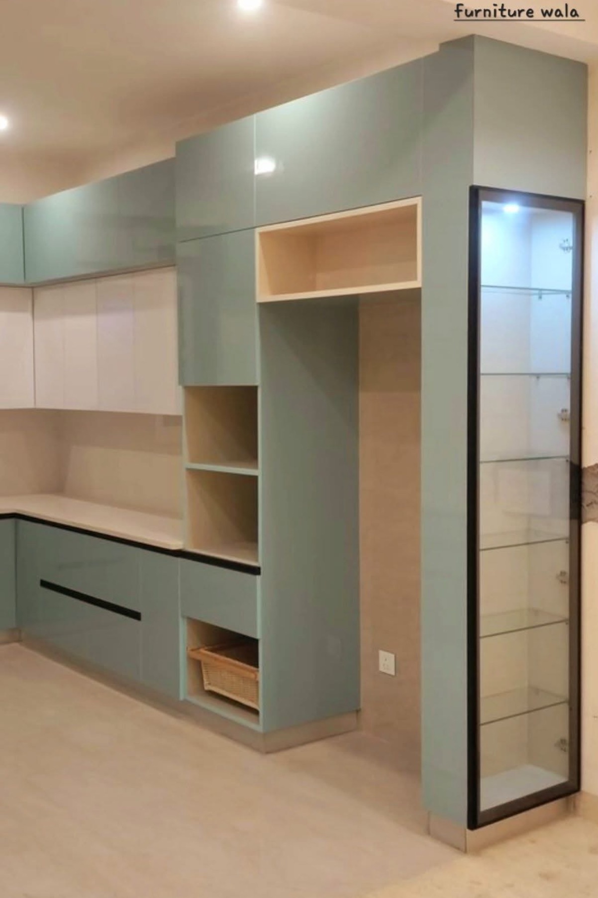 Nowoczesna kuchnia na wymiar z błękitnymi frontami i przeszkloną witryną z oświetleniem. W dolnej szafce widoczny koszyk. Jasne blaty i minimalistyczne uchwyty.