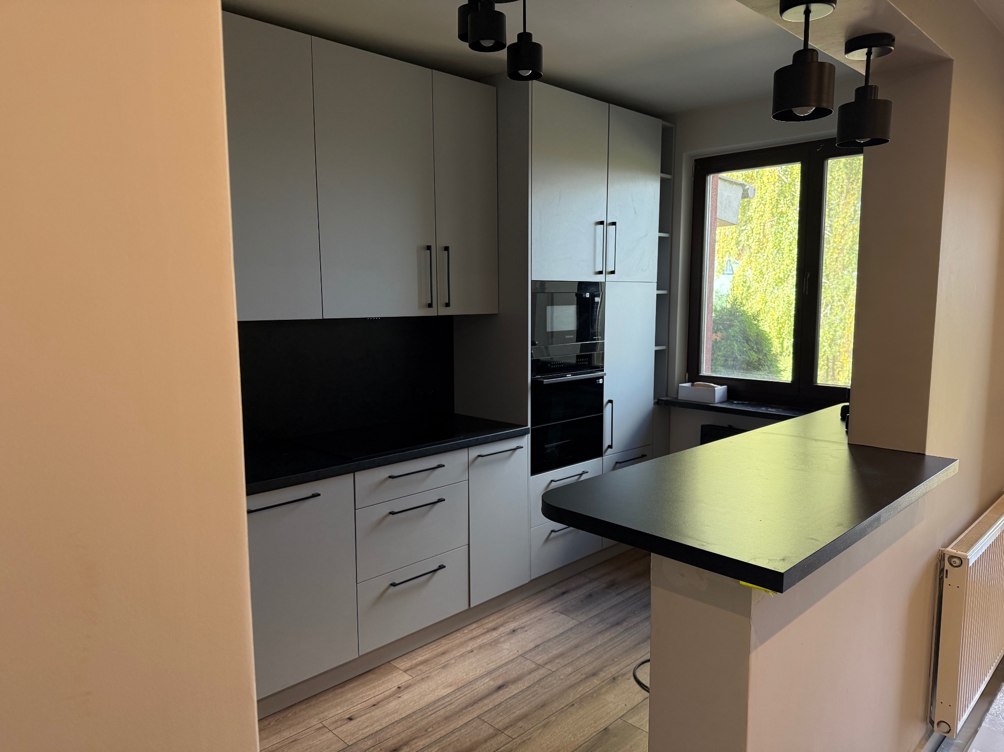 Jasna kuchnia z czarnym blatem i oświetleniem punktowym. Widok na okno z zielenią. Nowoczesny design z drewnianą podłogą i minimalistycznymi uchwytami.