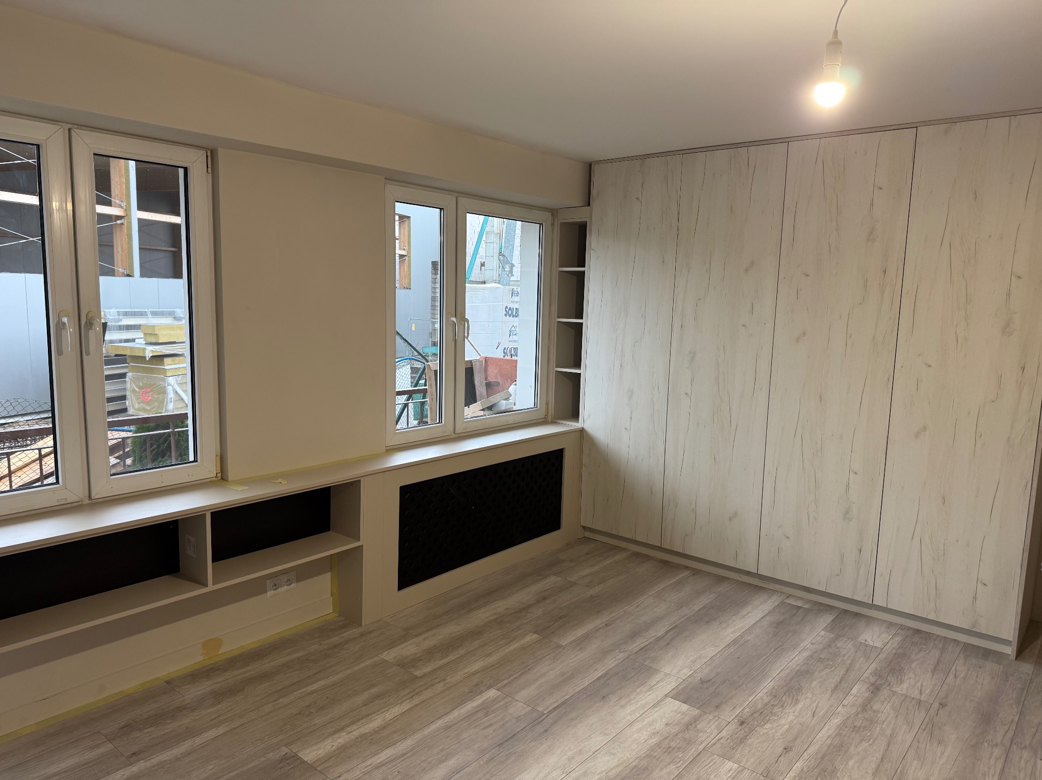 Jasne wnętrze po remoncie: drewniana podłoga, zabudowa okienna z półkami, grzejnik w obudowie i szafa wnękowa z frontami imitującymi drewno. Widok na zewnątrz przez okna.