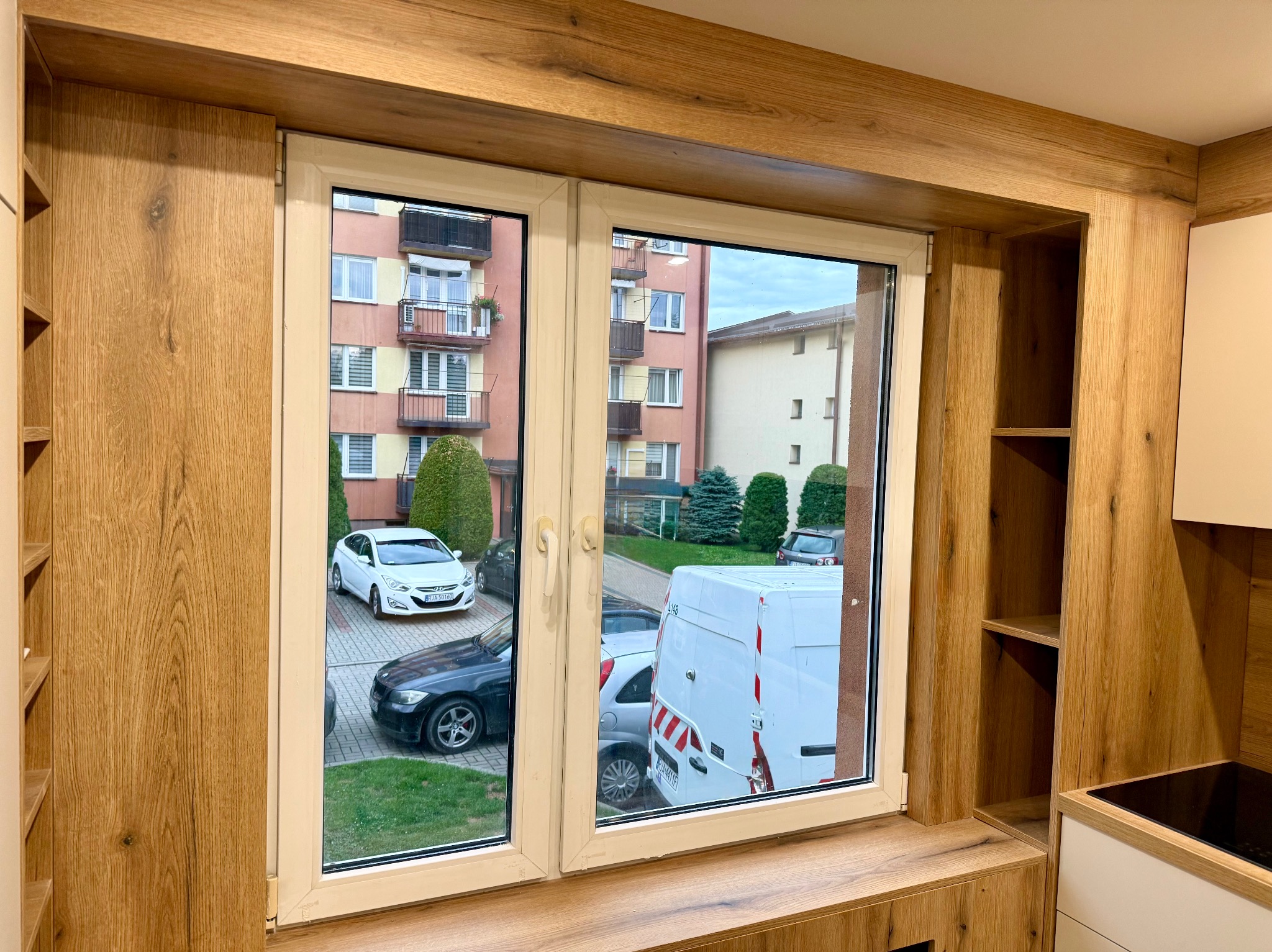 Okna PCV w drewnianej obudowie, widok na parking z samochodami. Zabudowa okienna z półkami, jasne drewno. Nowoczesny design.