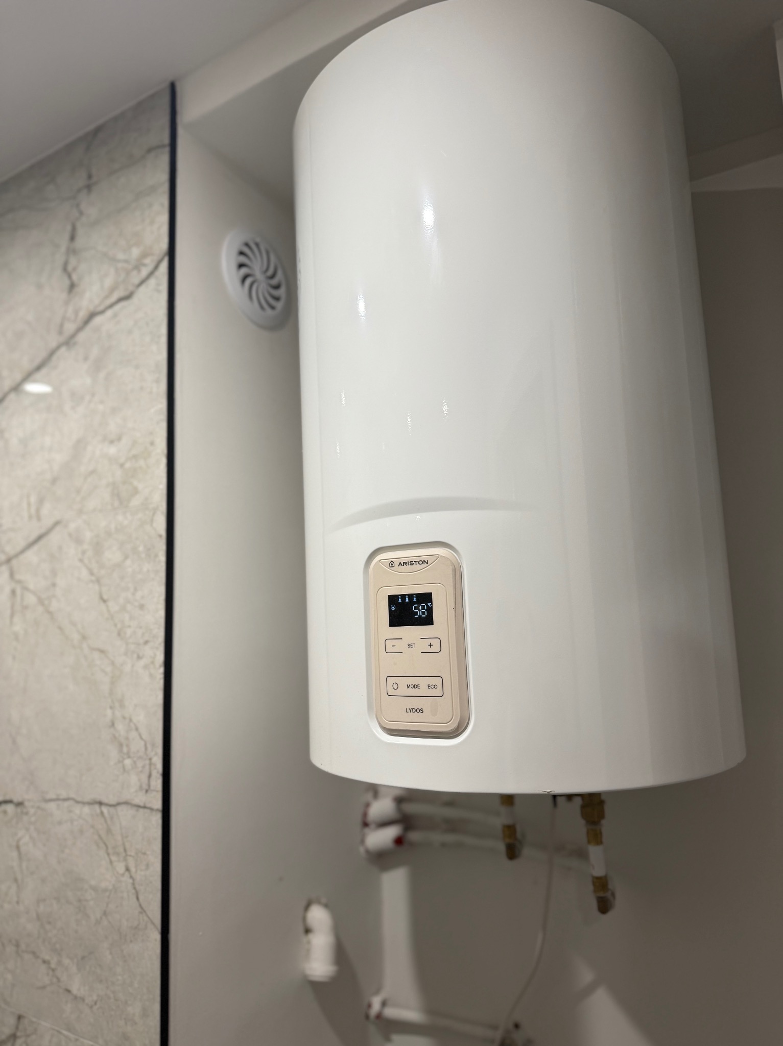 Biały, cylindryczny bojler Ariston Lydos z wyświetlaczem 58°C, zamontowany na ścianie w łazience z marmurową płytką. Widoczne rury doprowadzające wodę.