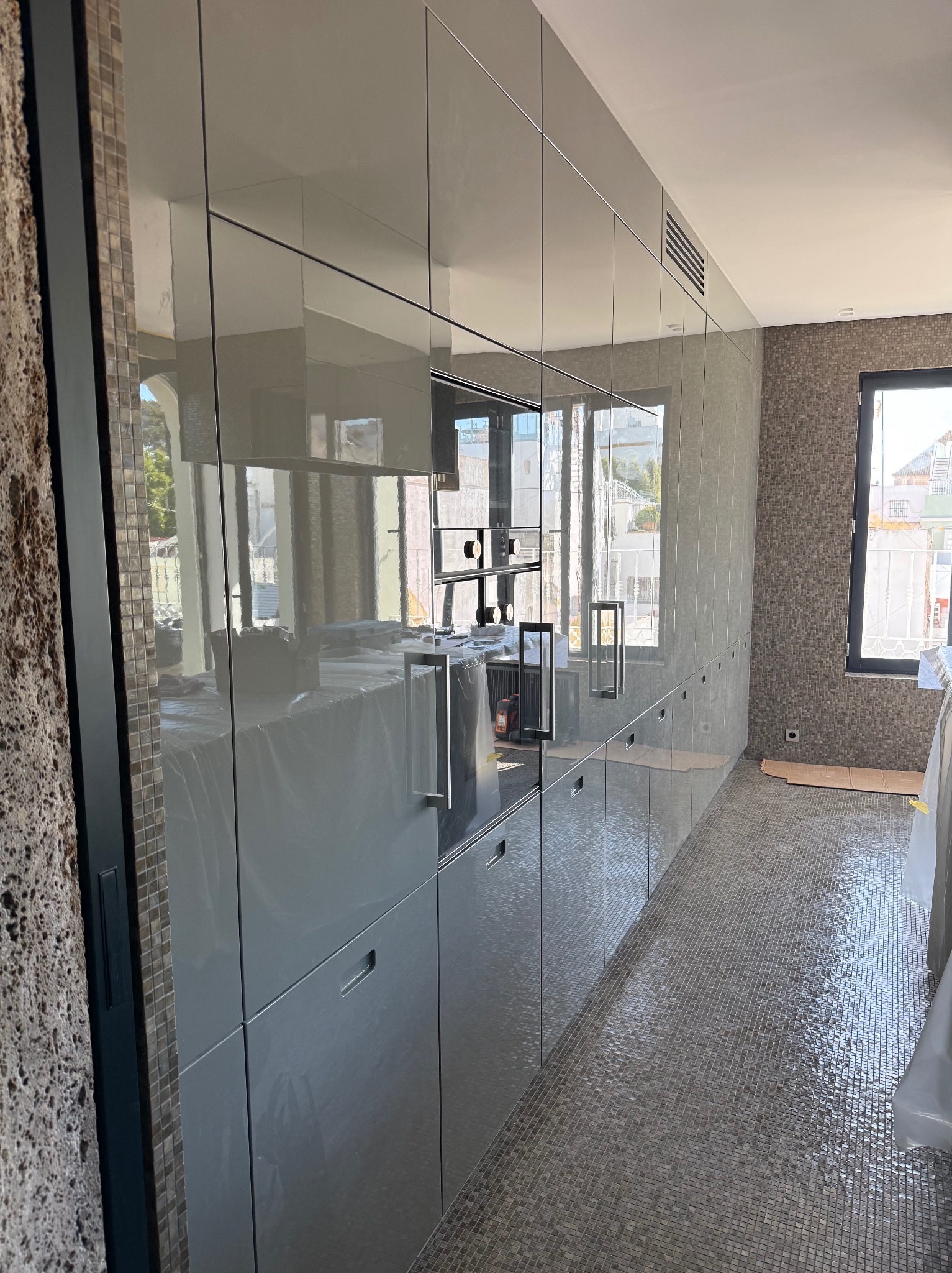 Nowoczesna kuchnia z połyskiem, szare szafki na całej ścianie, mozaika na podłodze i ścianie, okno z widokiem na zewnątrz. Część szafek zabezpieczona folią.