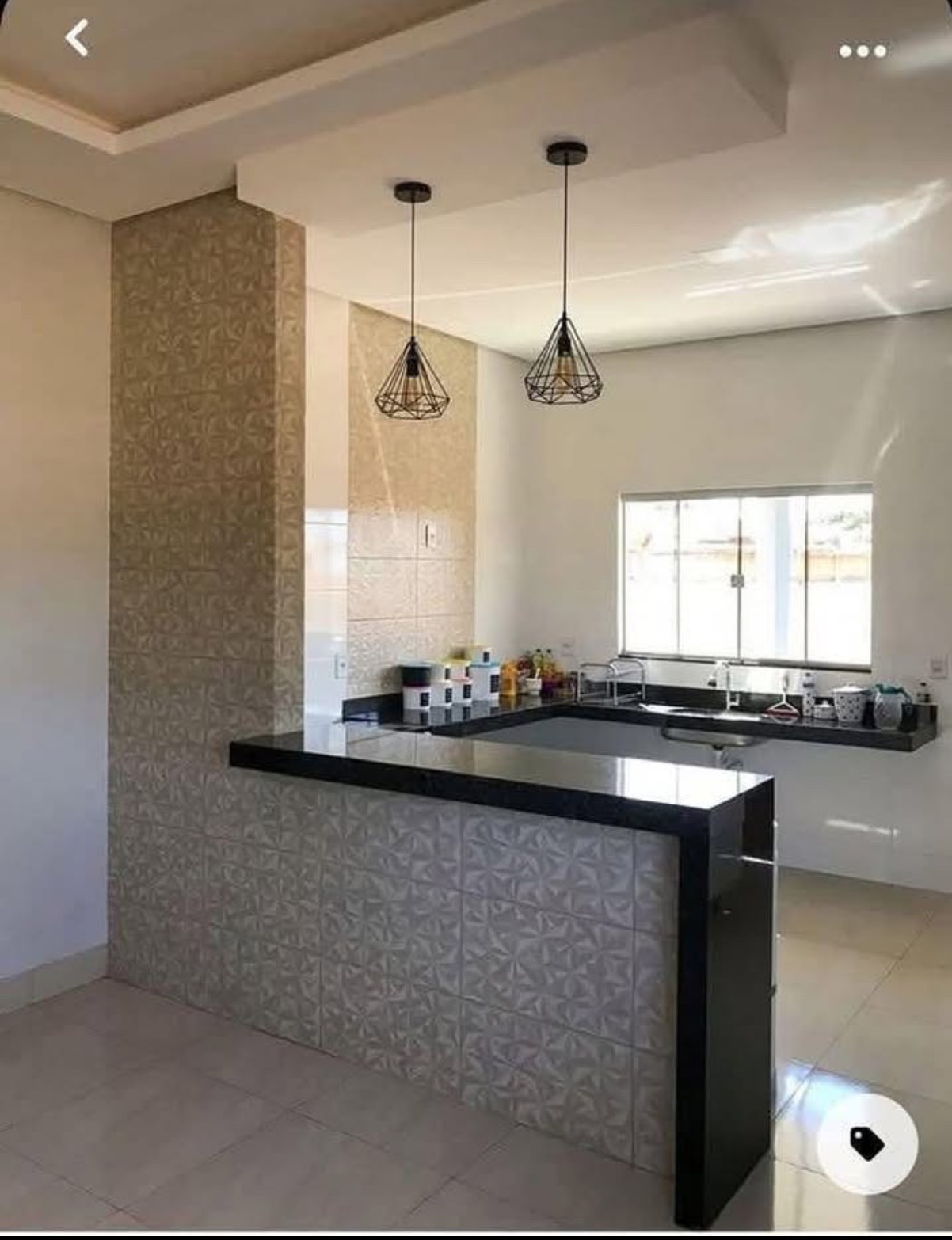 Wykończona kuchnia z wyspą wyłożoną wzorzystymi płytkami, czarny blat, nowoczesne lampy wiszące. Widok na okno i blat roboczy. Jasne wnętrze.