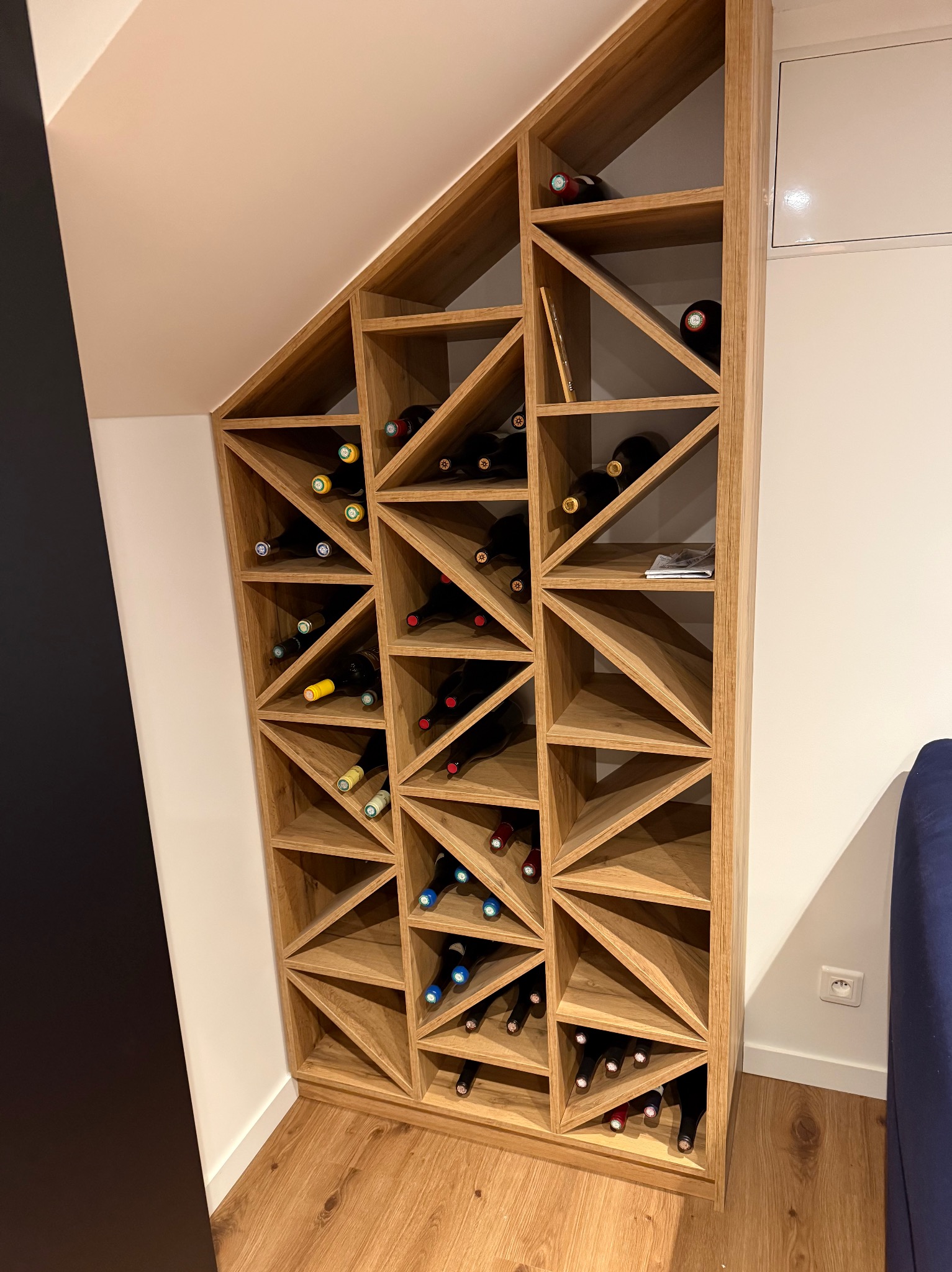Drewniany regał na wino, wkomponowany w skos pomieszczenia, wypełniony butelkami wina z kolorowymi etykietami. Widoczna podłoga z paneli i fragment niebieskiej sofy.
