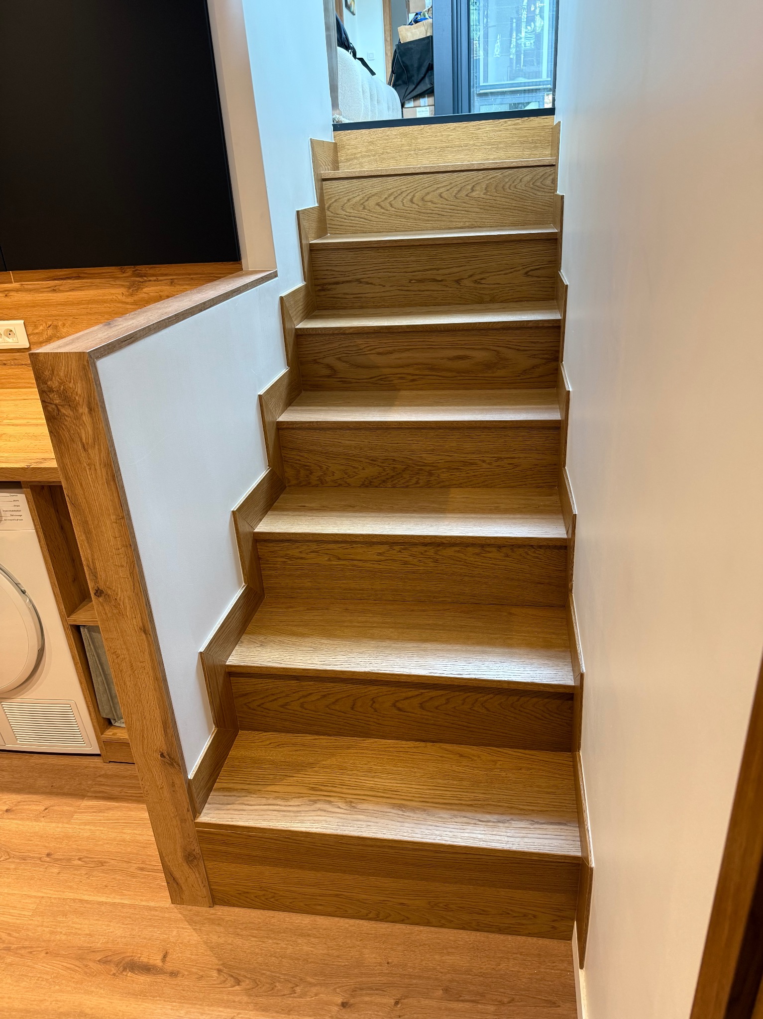 Drewniane schody z białymi ścianami, widok z dołu. Naturalne oświetlenie podkreśla strukturę drewna. Fragment zabudowy kuchennej z pralką w tle.