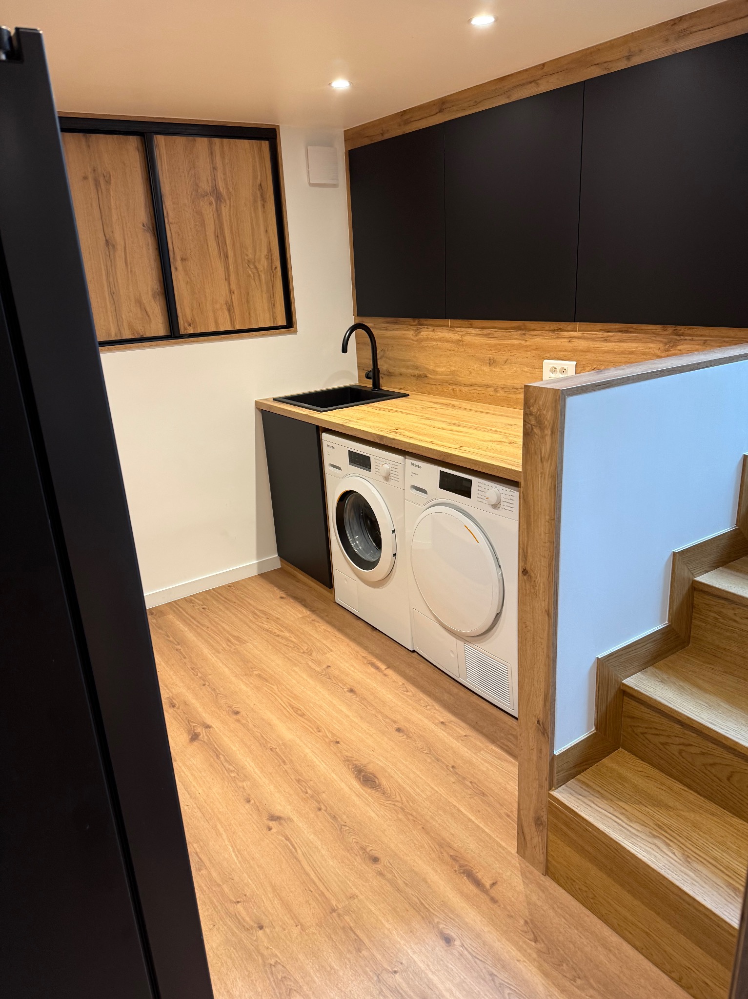Nowoczesna pralnia: pralka i suszarka Miele pod blatem z czarnym zlewem, czarne szafki wiszące, drewniane wykończenia i schody. Minimalistyczny design.