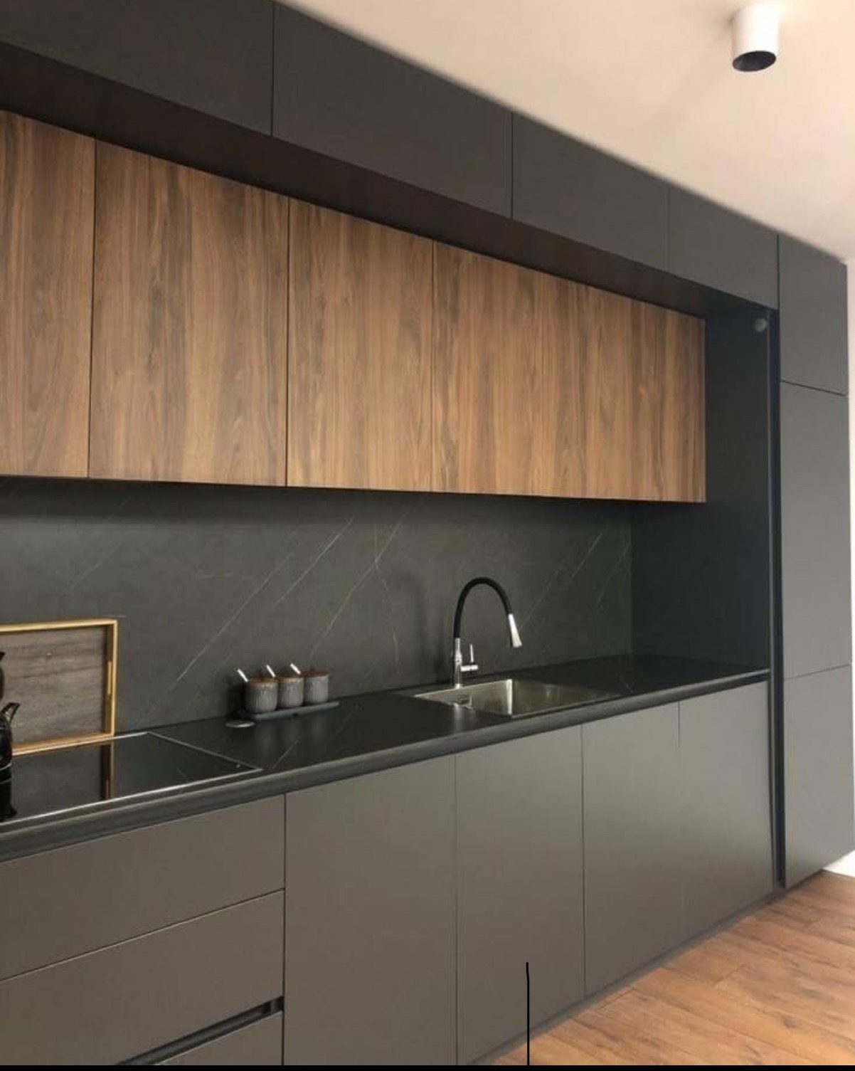 Nowoczesna kuchnia z szarymi szafkami i drewnianymi górnymi szafkami. Czarny blat i backsplash z marmurowym wzorem. Minimalistyczny design, oświetlenie punktowe.