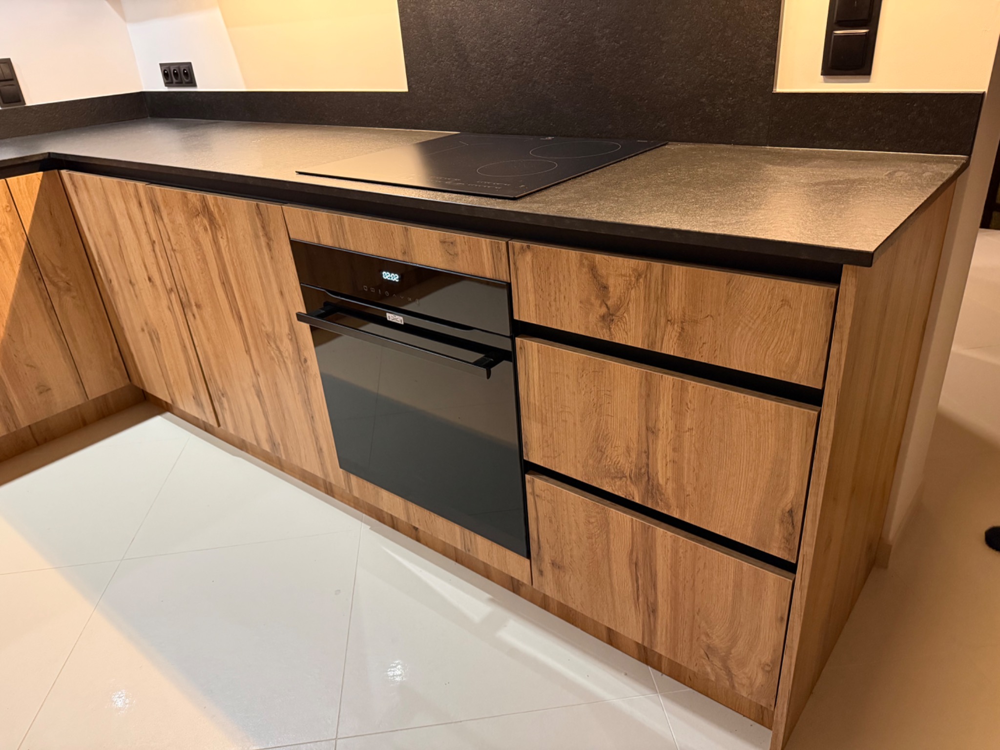 Nowoczesna zabudowa kuchenna z drewnianymi frontami, czarnym blatem i płytą indukcyjną. Widoczny piekarnik w zabudowie. Białe płytki na podłodze.