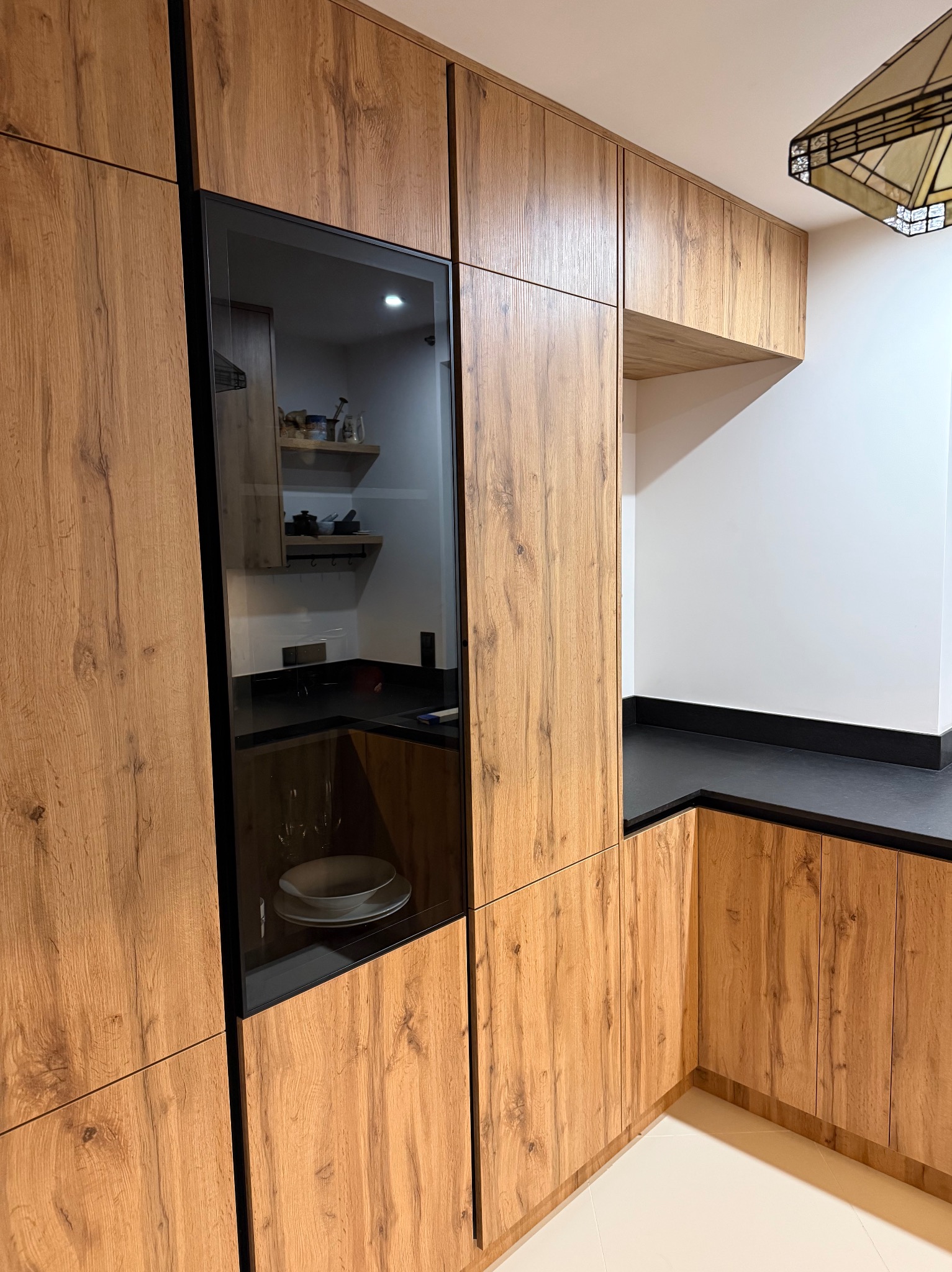 Kuchnia na wymiar z drewnianymi frontami i czarnym blatem. W tle widoczny fragment oświetlenia sufitowego. Fronty szafek z wyraźnym rysunkiem drewna.