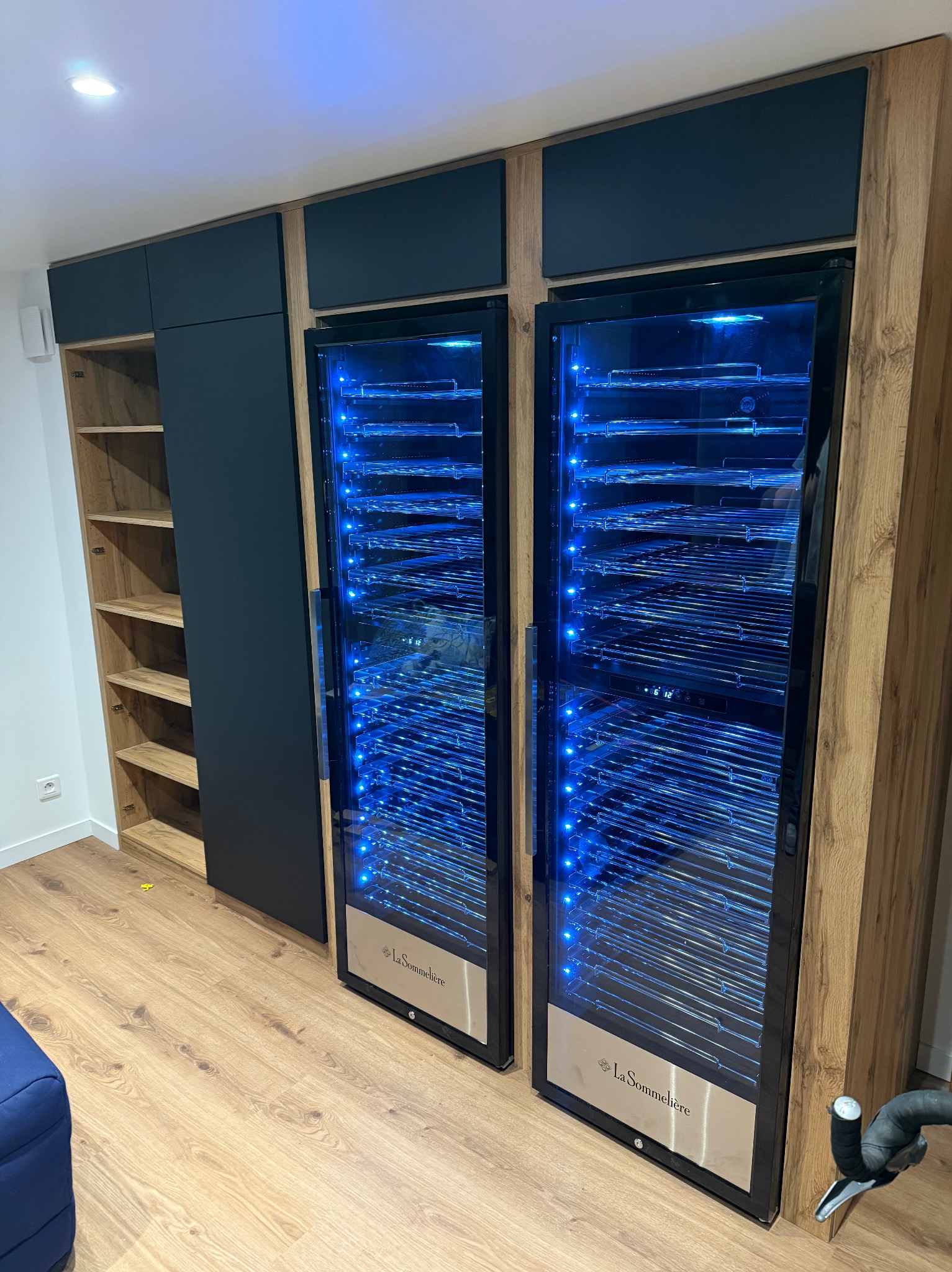 Dwie szafy chłodnicze na wino La Sommeliere z niebieskim oświetleniem LED, wkomponowane w zabudowę meblową z drewnianymi i czarnymi elementami.