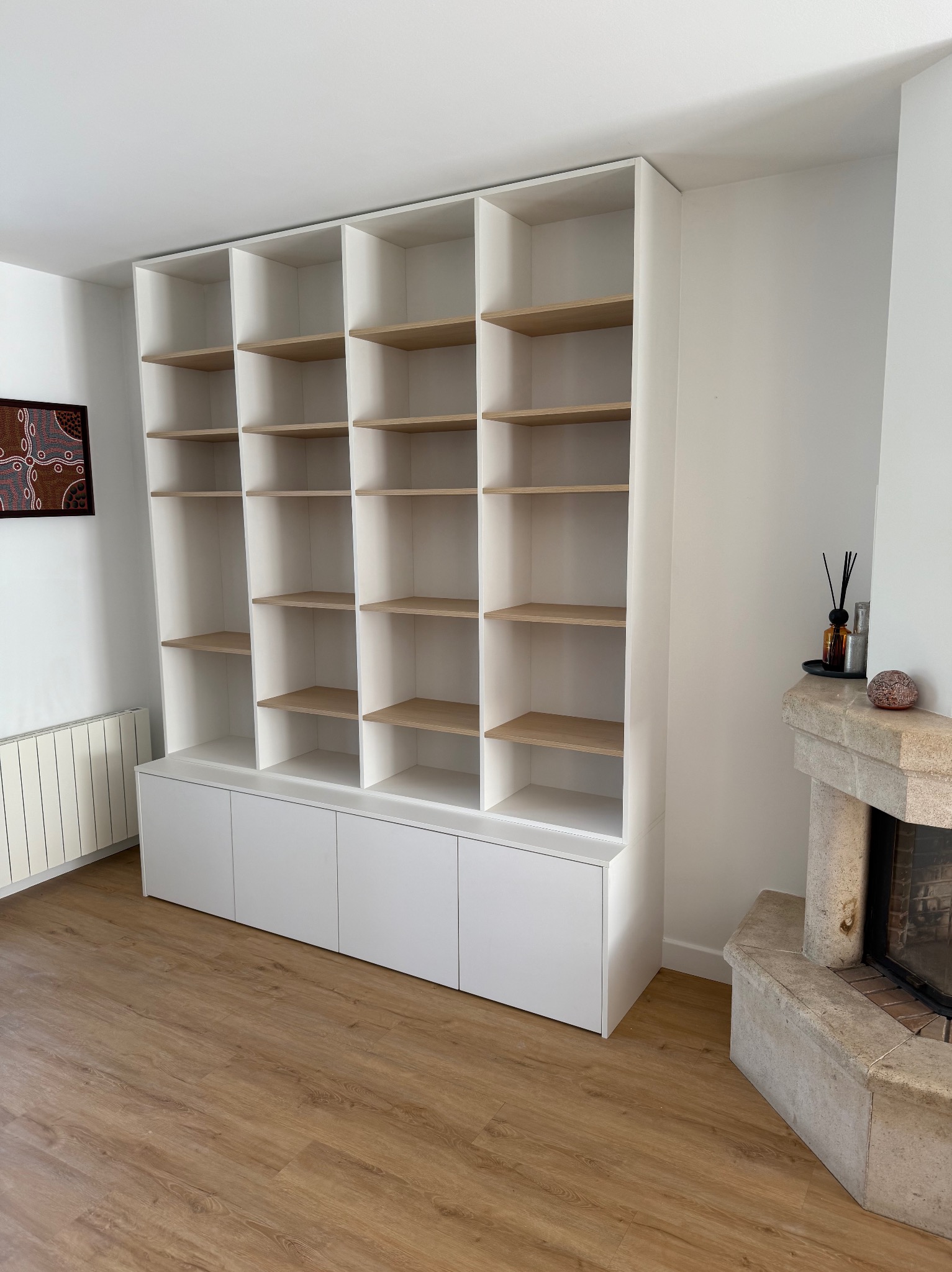 Biały regał na wymiar z jasnymi półkami, wbudowany w ścianę, z dolnymi szafkami. Obok kominek z kamienia i dekoracje. Minimalistyczny design, jasne wnętrze.