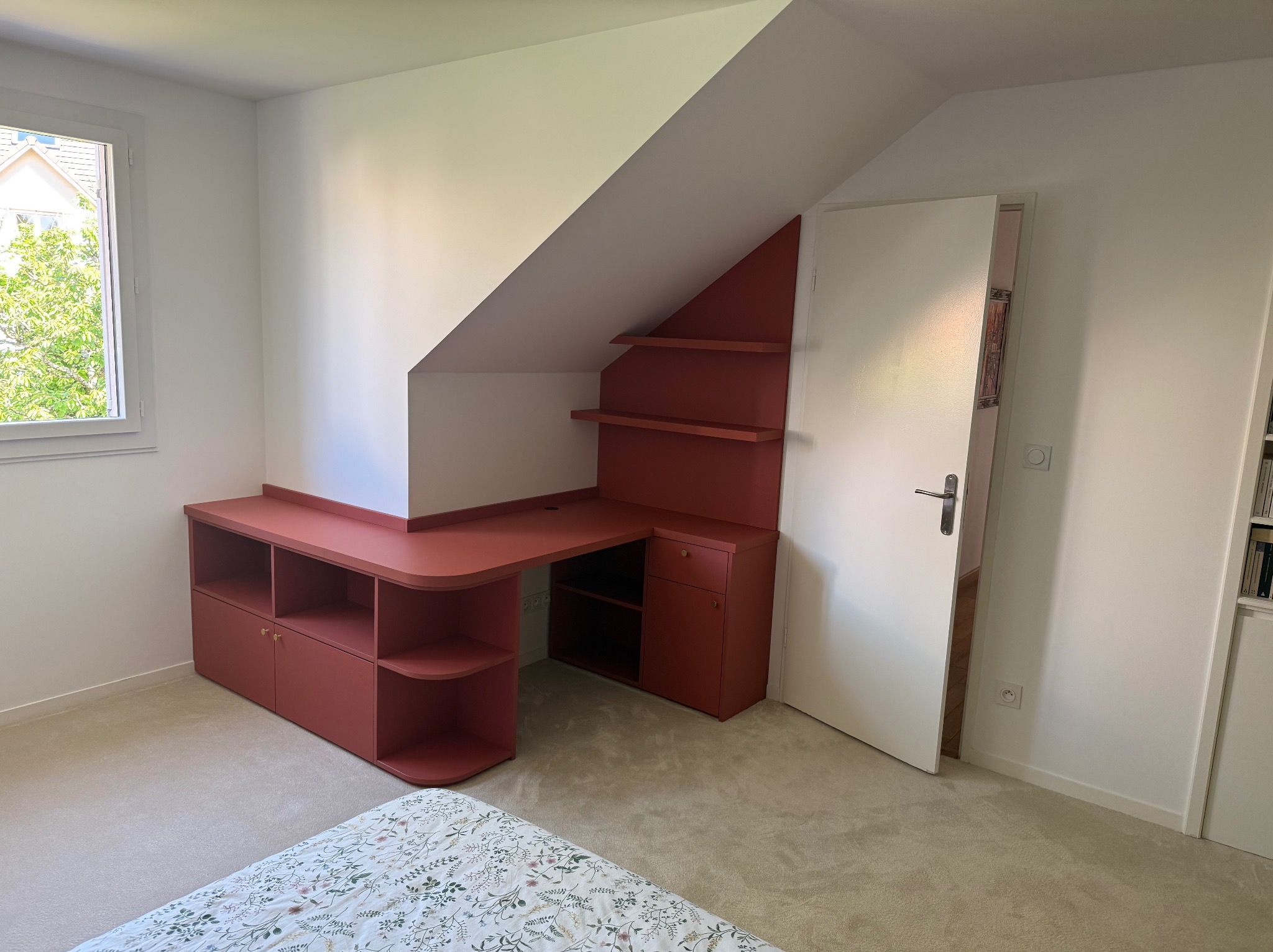 Czerwone biurko na wymiar w pokoju ze skosami, z półkami i szafkami, dopasowane do przestrzeni pod skosem. Widok na okno i fragment łóżka.