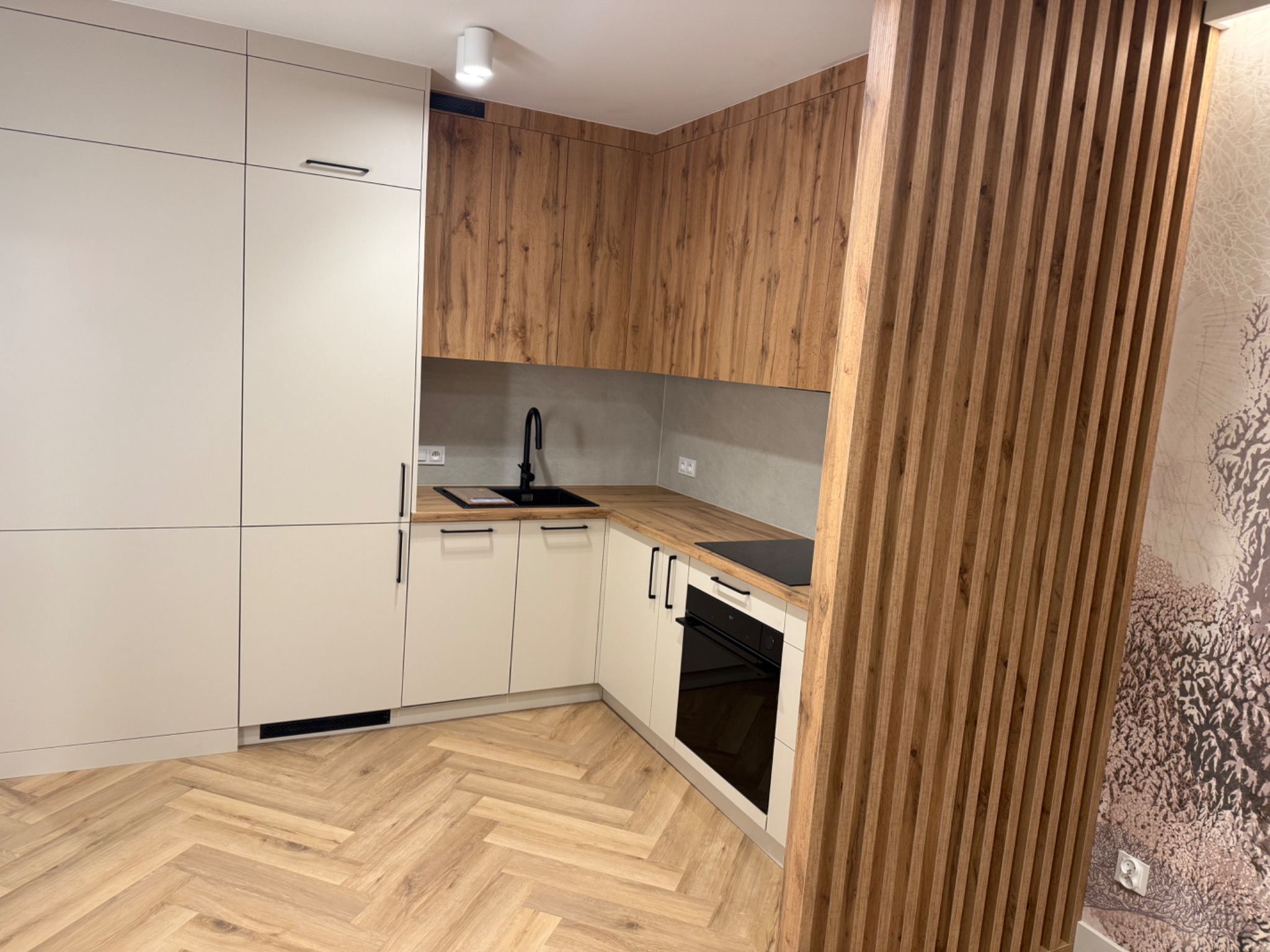 Nowoczesna kuchnia z białymi szafkami i drewnianymi akcentami, parkiet w jodełkę, pionowe lamele dekoracyjne i czarna armatura. Minimalistyczny design.