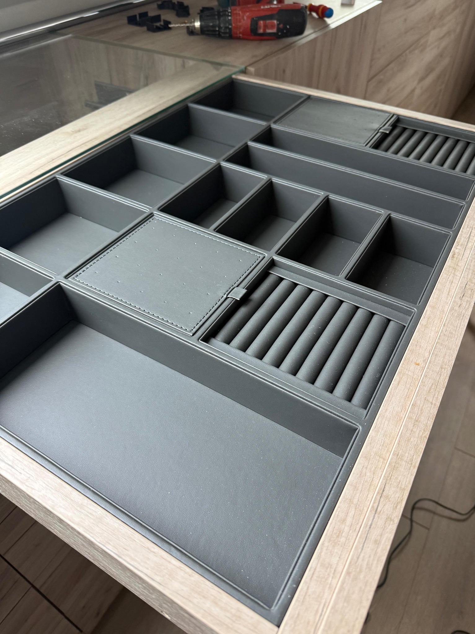 Szary organizer z przegródkami na biżuterię w szufladzie z drewnianym wykończeniem, widoczny w górnym rogu wkrętak i akcesoria montażowe.