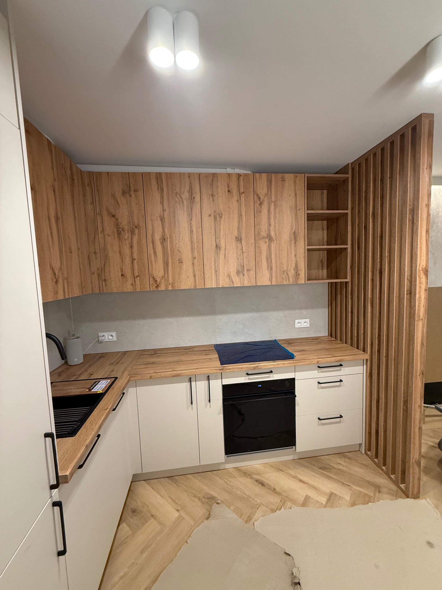 Nowoczesna kuchnia z drewnianymi szafkami i białymi frontami, wykończona drewnianą ścianką działową. Parkiet w jodełkę, czarny zlew i piekarnik.