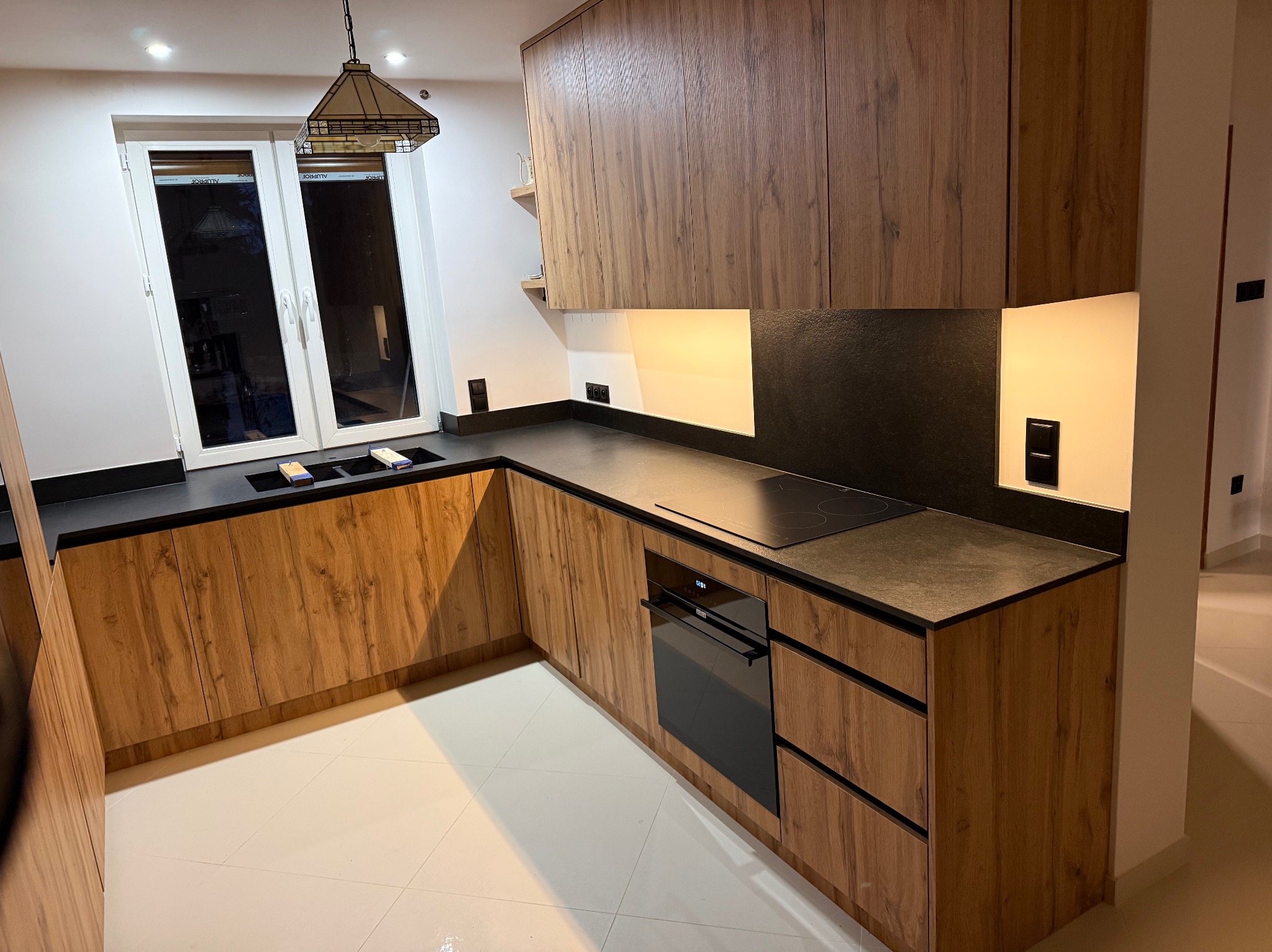 Nowoczesna kuchnia na wymiar z drewnianymi frontami i czarnym blatem w Jarosławiu. Widoczna płyta indukcyjna i piekarnik. Minimalistyczny design, ciepłe oświetlenie.