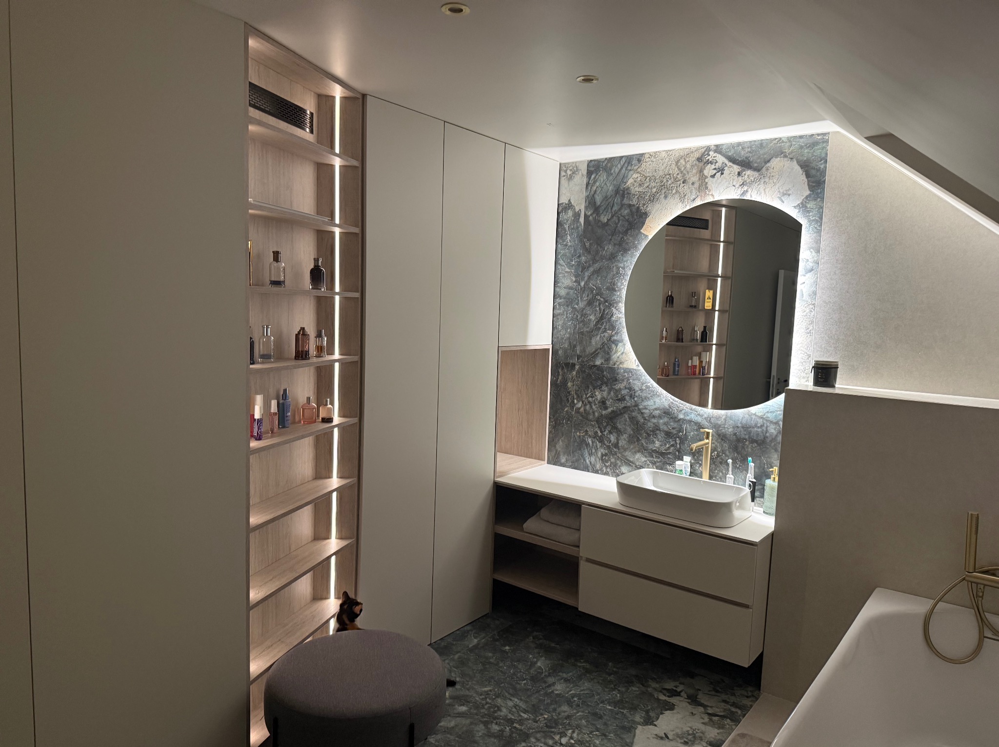 Nowoczesna łazienka na poddaszu z szafkami na wymiar, okrągłym lustrem z podświetleniem LED i marmurową posadzką. Widoczny fragment wanny i pufa.